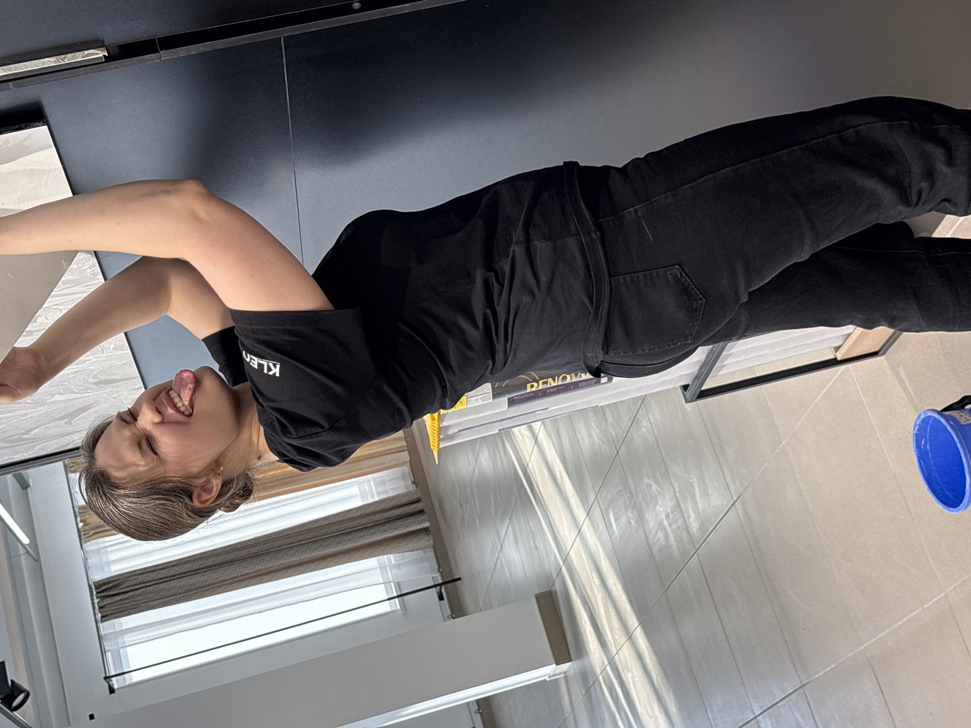 A woman with graying hair is sticking her tongue out and lying on a black surface, possibly a table or counter. She is wearing a black t-shirt with the word "KLEEN" printed on it and black jeans. The background shows a window with blinds, a white cabinet, and tiled flooring. A blue bucket is visible in the bottom right corner. The scene appears to be indoors. The exact location in Chaikovsky, Russia, is indeterminable from the image.