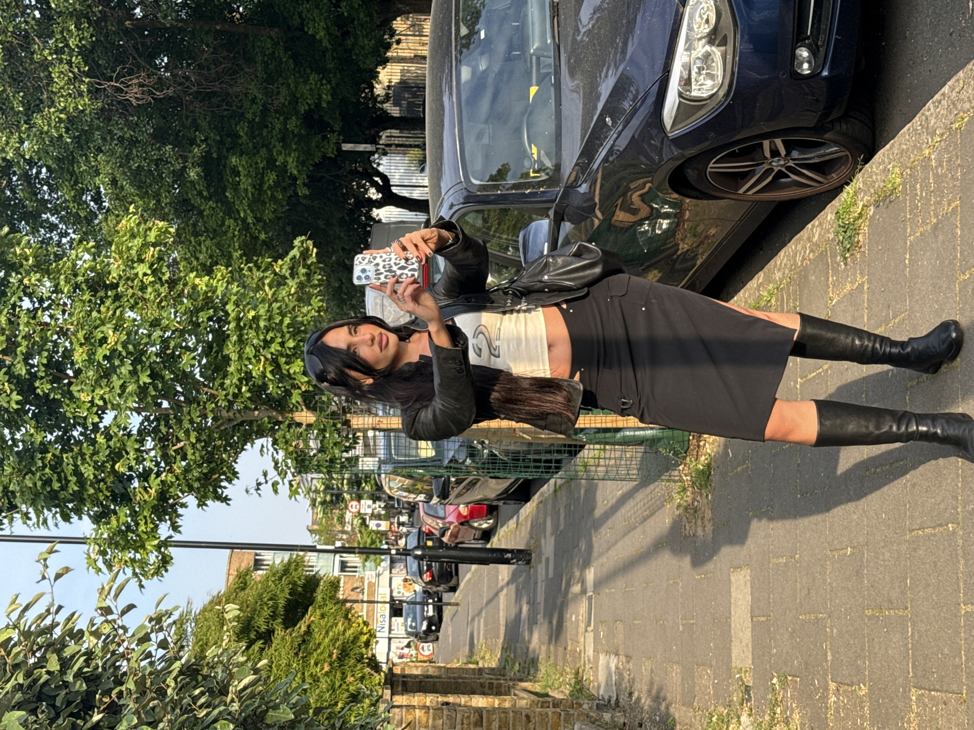 A woman stands on a London sidewalk next to a parked car, taking a photo with her phone. The location is a residential street lined with houses, trees, parked cars, and street signs. The woman is wearing a black leather jacket, a white crop top, a black skirt, and knee-high black boots. She's holding a phone with a patterned case. Additional cars are parked down the street, and greenery is visible on both sides of the road. The sidewalk is paved with interlocking bricks. The weather appears sunny and bright.