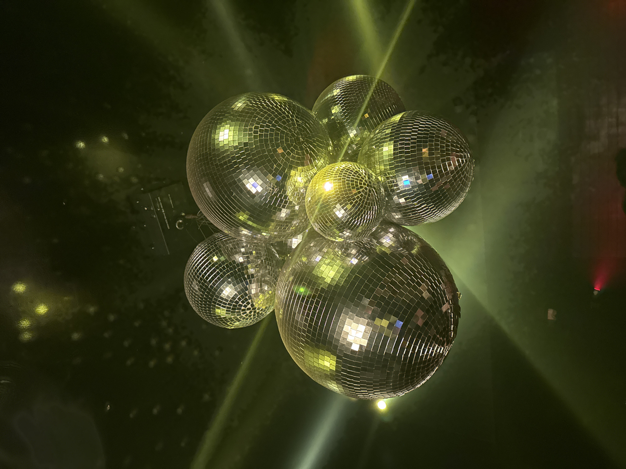 The image shows a cluster of disco balls hanging from a ceiling, illuminated by green and yellow lights, suggesting a nightlife venue. The disco balls are spherical and covered in small, reflective mirrored squares. Beams of colored light cut across the dark, smoky atmosphere. In the lower right corner, a faint red light is visible, possibly from another light fixture or stage lighting. No people are clearly identifiable, though some dark shapes at the very bottom suggest a crowd. The scene is dark and moody, with the disco balls creating a festive, reflective display. The image's composition is tight on the disco balls, with the background providing a sense of the environment without much detail. The location is not specified as Paris, France.