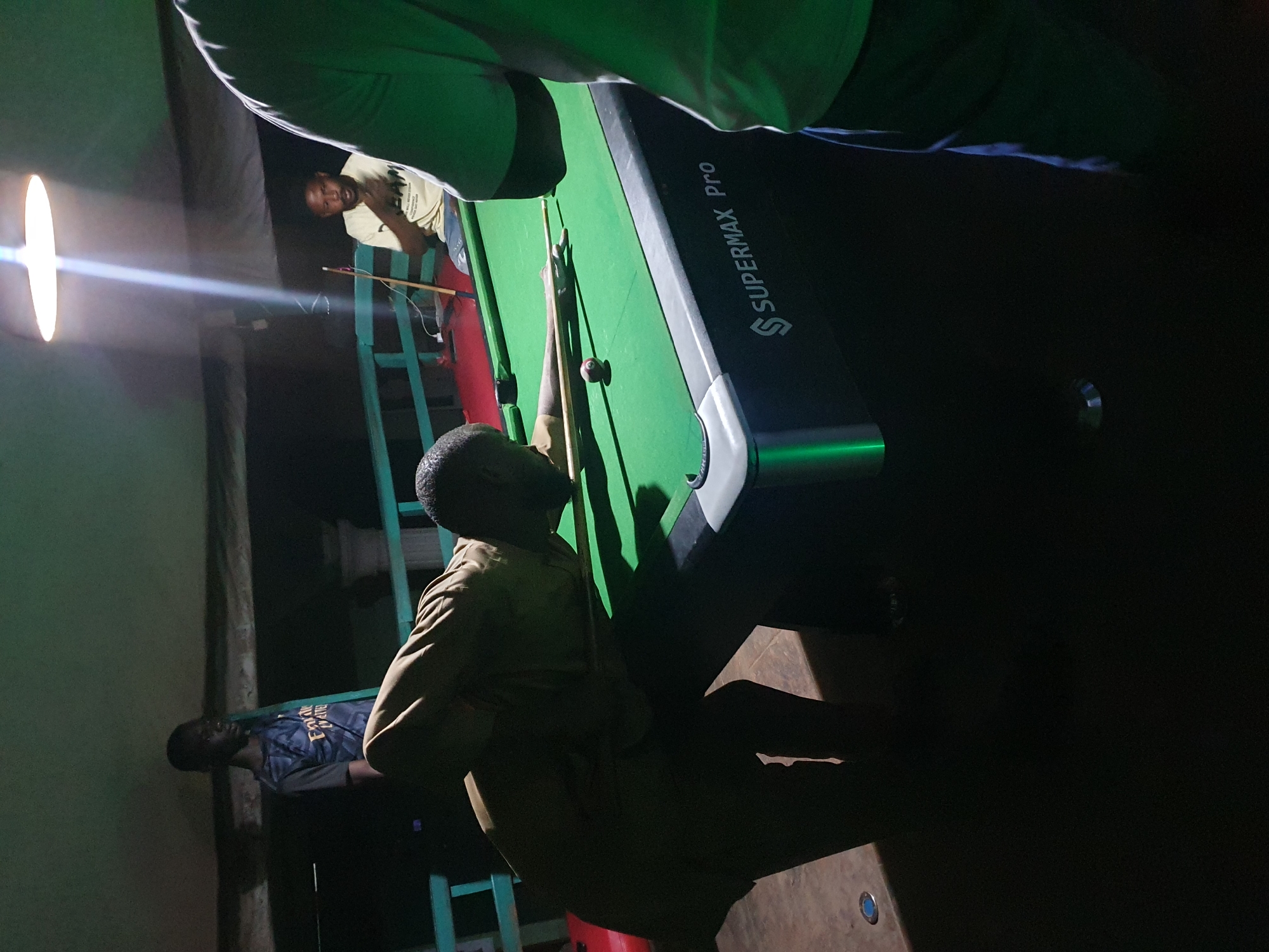 The image depicts an indoor scene at night, likely in Lokoja, Nigeria, featuring a pool table and several individuals. The primary focus is on a person in the foreground, crouched over the green pool table, in the process of playing billiards. They are wearing a light brown or tan outfit and their face is obscured by their posture. To the right, another person is partially visible, wearing a light-colored t-shirt and dark pants, seemingly observing the game. In the background, a third individual stands, wearing a dark t-shirt with some lettering, possibly a sports jersey. A bright overhead light illuminates the scene, casting distinct shadows. The pool table itself is dark with a "SUPERMAX Pro" logo visible on its side. The environment appears to be somewhat dimly lit beyond the immediate playing area, with a hint of a wooden railing or structure in the background.