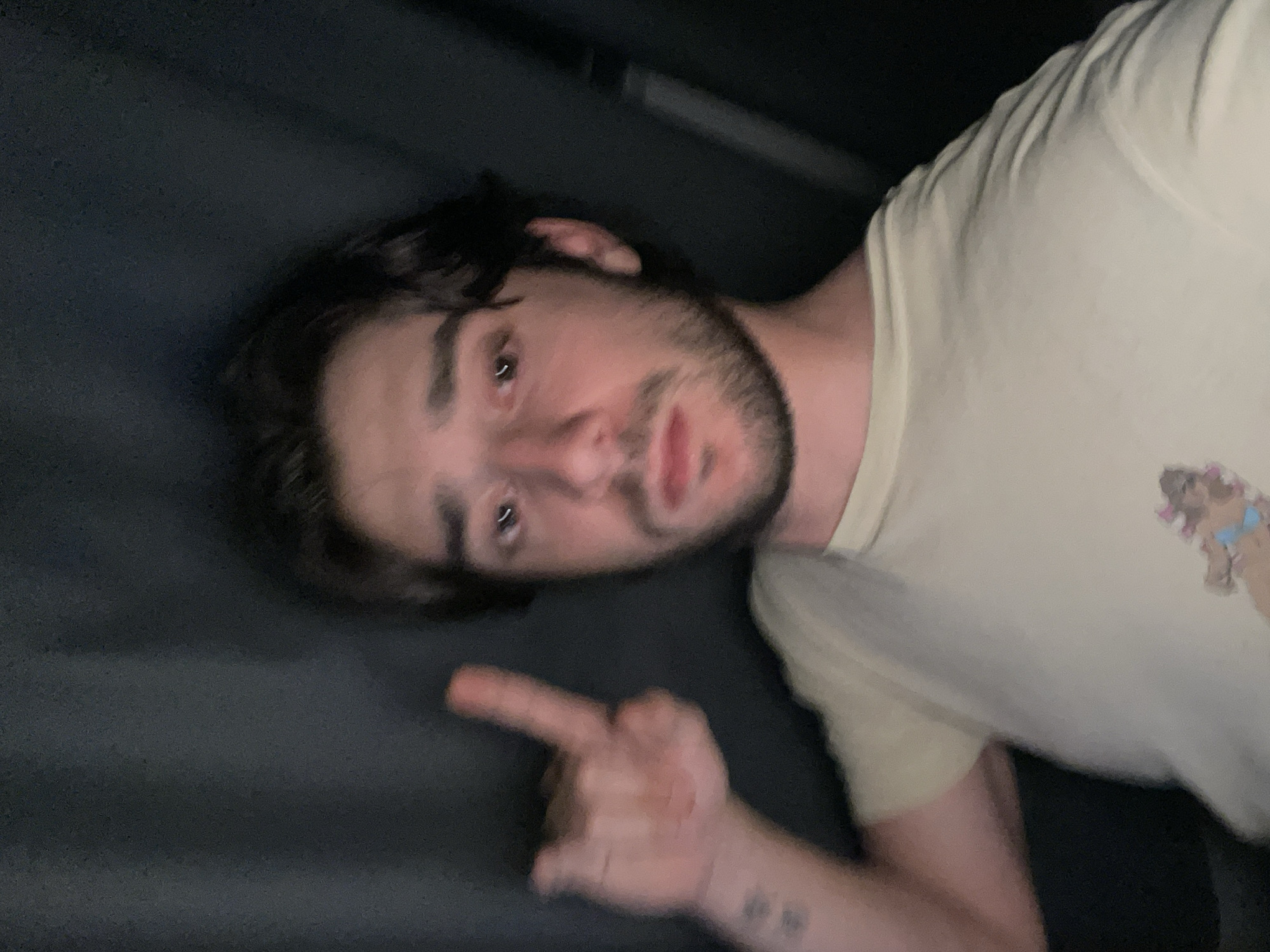 In Huixquilucan de Degollado, Mexico, a young person with dark hair and a beard is taking a selfie. They are wearing a light-colored t-shirt with a graphic print. Their left hand is raised, with the index finger pointing upwards. The background is dark and appears to be a curtain or fabric. The lighting is dim, and the image is slightly blurry.