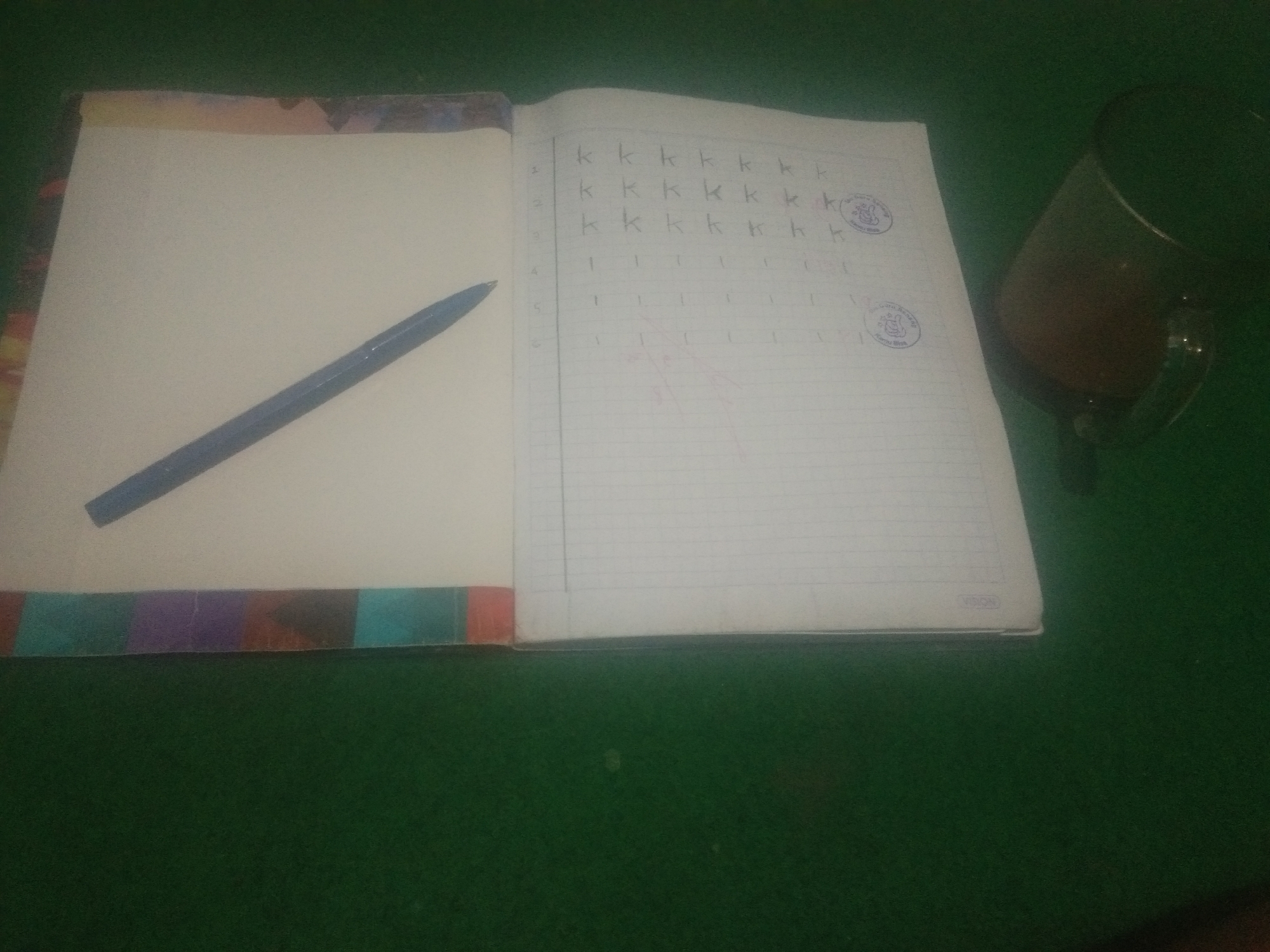 An open notebook lies on a green surface, a blue pen resting on the left page. The right page of the notebook is lined, with rows of the letter "k" handwritten in pencil. A glass filled with a dark liquid sits to the right of the notebook. The image is taken from a high angle. The location of Bulusari, Indonesia, is not discernible from the visual information in the image.
