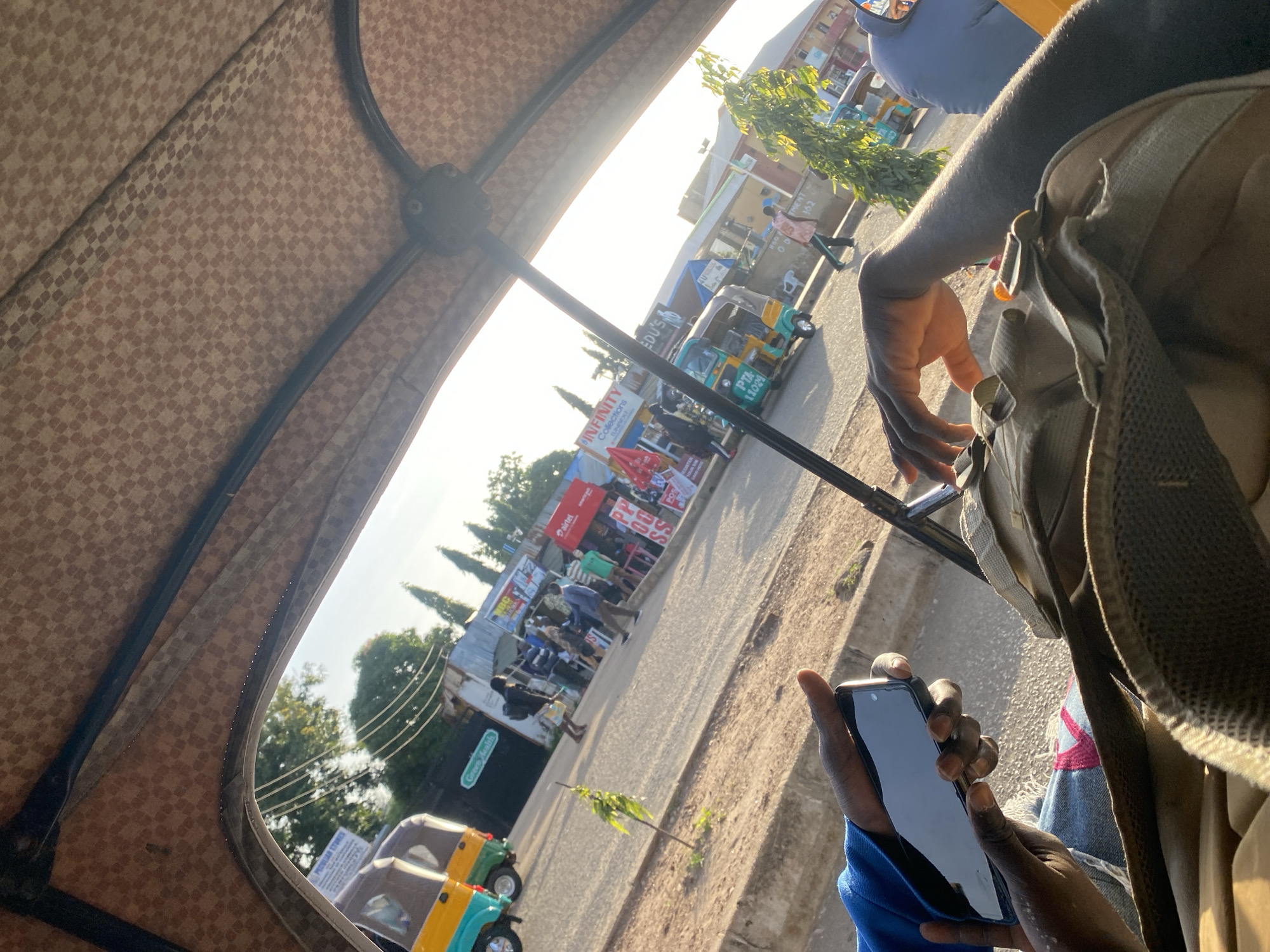 The image shows a first-person view from inside a motorized tricycle, commonly known as a "Keke Napep," in Jos, Nigeria. The tricycle's patterned canvas roof is visible at the top. In the foreground, a person's hands are holding a black smartphone, and a backpack rests on their lap.
Outside the tricycle, a street scene unfolds. Several other Keke Napeps are present, along with pedestrians. Buildings line the street, some of which appear to be shops with signs indicating businesses. One sign visible is for "Green Health." The overall lighting suggests it is daytime, possibly late afternoon, with the sun casting long shadows and a warm glow. The environment appears to be a busy urban area.