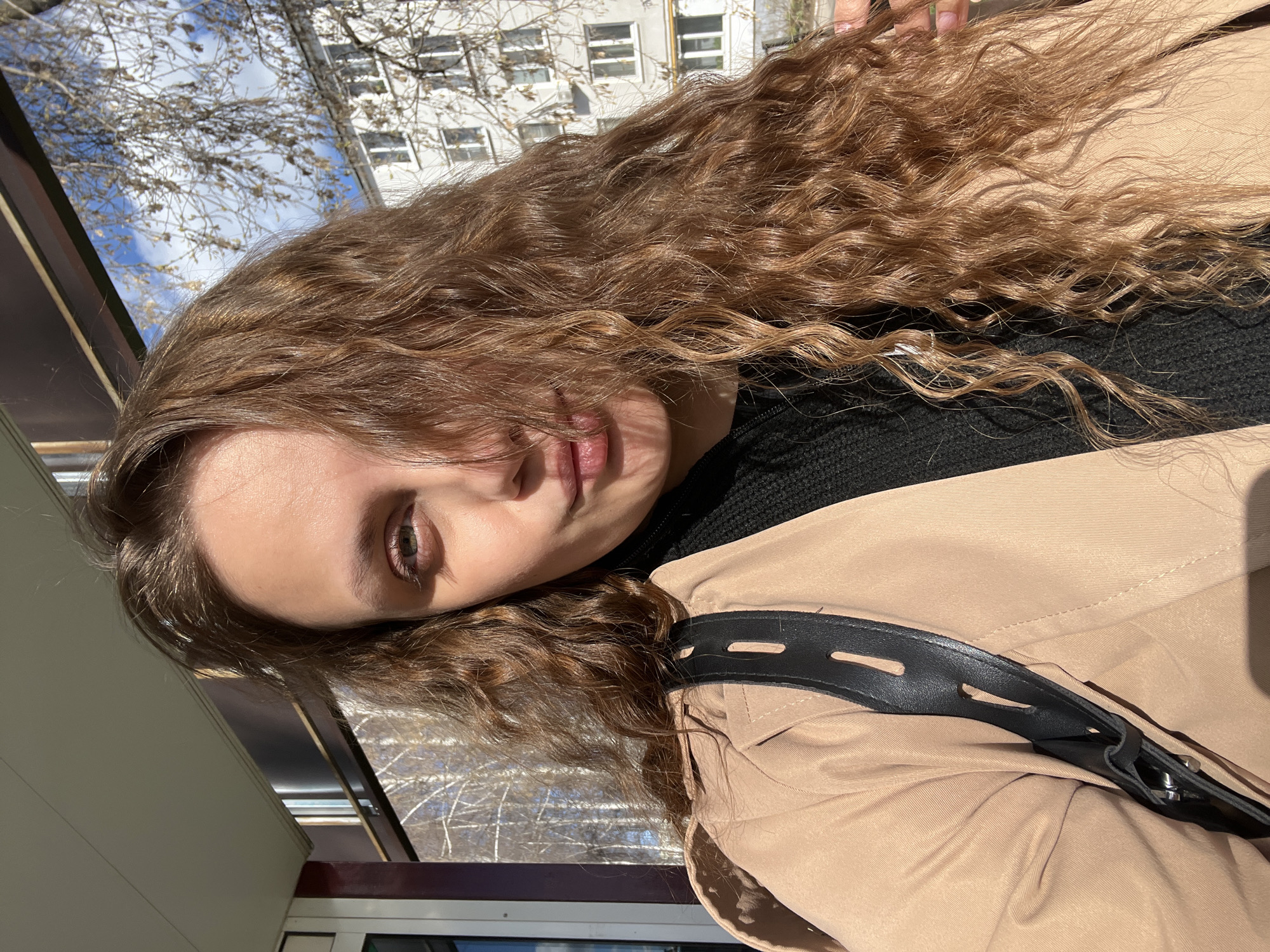A young woman with long, curly brown hair is positioned in front of a window with a cityscape visible in the background. The woman is wearing a black top underneath a tan jacket with a black leather strap. The setting appears to be in Chaikovsky, Russia, inferred from building styles and window structure. The woman is indoors, possibly on a train car or in an enclosed area with windows facing outwards. Sunlight illuminates the scene.
