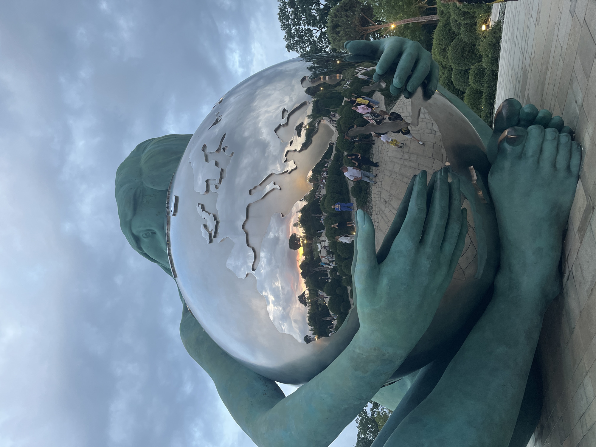 The image depicts a monumental outdoor sculpture of a large, stylized figure cradling a metallic sphere. The sculpture is a deep greenish-blue color, with a textured surface. The sphere it holds is highly reflective, showing a distorted image of the surrounding environment, including a sunset with orange and pink hues in the clouds, as well as trees and bushes. Several people are visible in the reflected image, appearing to be walking in a park-like setting with paved pathways and manicured greenery. The sculpture itself appears to be made of a solid material, possibly bronze or patinated metal. The overall scene suggests a public art installation in a park or urban space. Based on the distinctive sculpture, it is identified as the monument "The Birth of a New Man" by Zurab Tsereteli, located in the city of Krasnodar, Russia.
