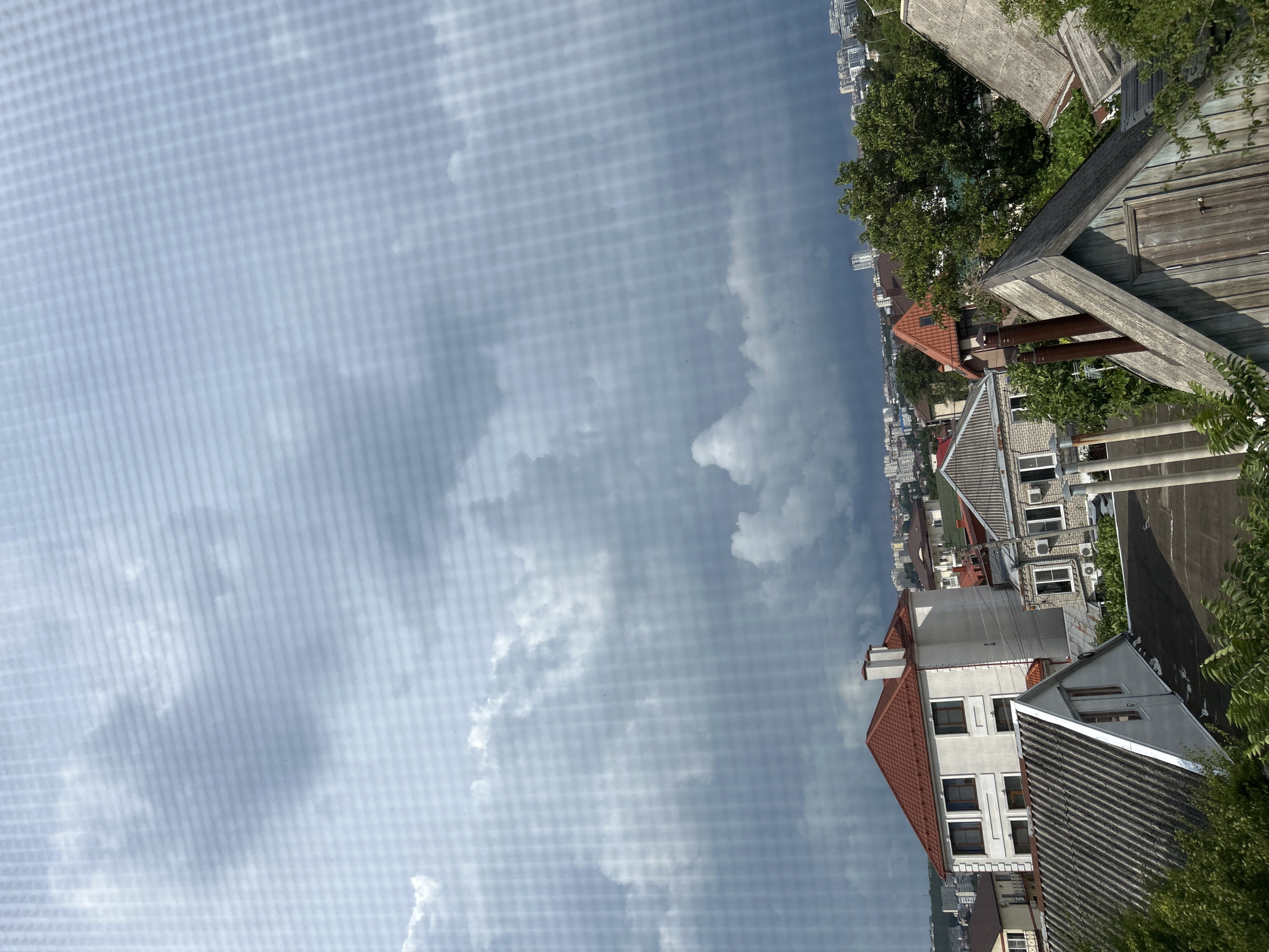 This image shows a residential area in Gelendzhik, Russia, under a cloudy sky. The foreground features several rooftops, including one with a grey, corrugated material and another with a wooden facade and a small door. A white building with a red roof, featuring multiple windows, is prominent. Further in the distance, more houses and apartment buildings are visible, along with lush green trees. The sky is filled with dark, dramatic clouds, suggesting an impending storm. The perspective is from an elevated viewpoint, looking down and across the urban landscape. No people are visible in the image.