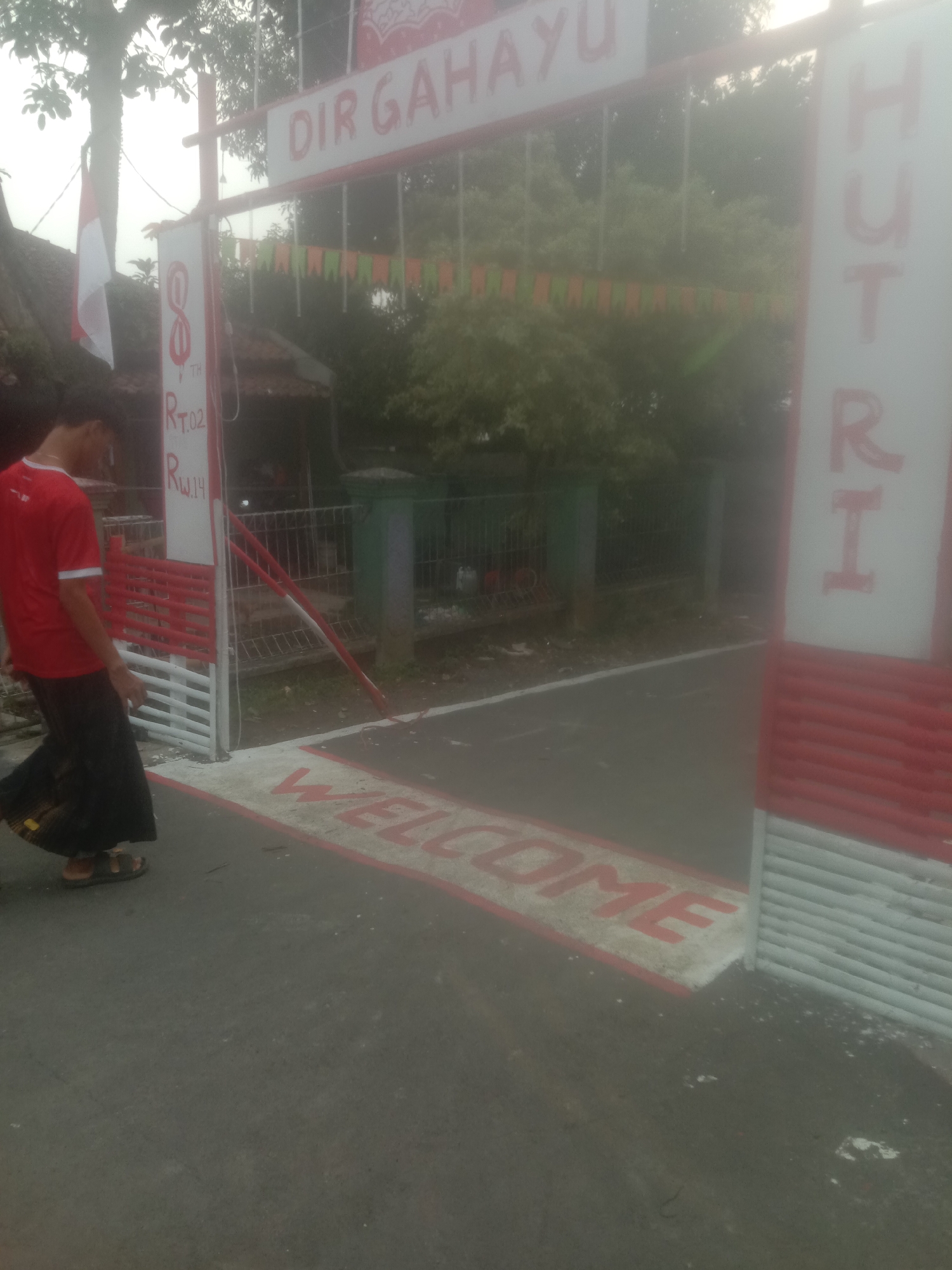 The image depicts an entrance gate decorated for a celebration. The gate features red and white striped pillars and horizontal bars. A banner at the top reads "DIR GAHAYU," and a vertical banner on the right side displays "HUTRI." Below the main banner, a string of green and orange flags is visible. A person wearing a red shirt and dark pants is walking through the gate. On the ground, the word "WELCOME" is painted in red letters within a white border. The scene appears to be in Bojong, Indonesia, as indicated by the Indonesian text on the banners. The environment is somewhat hazy, suggesting possible mist or fog.