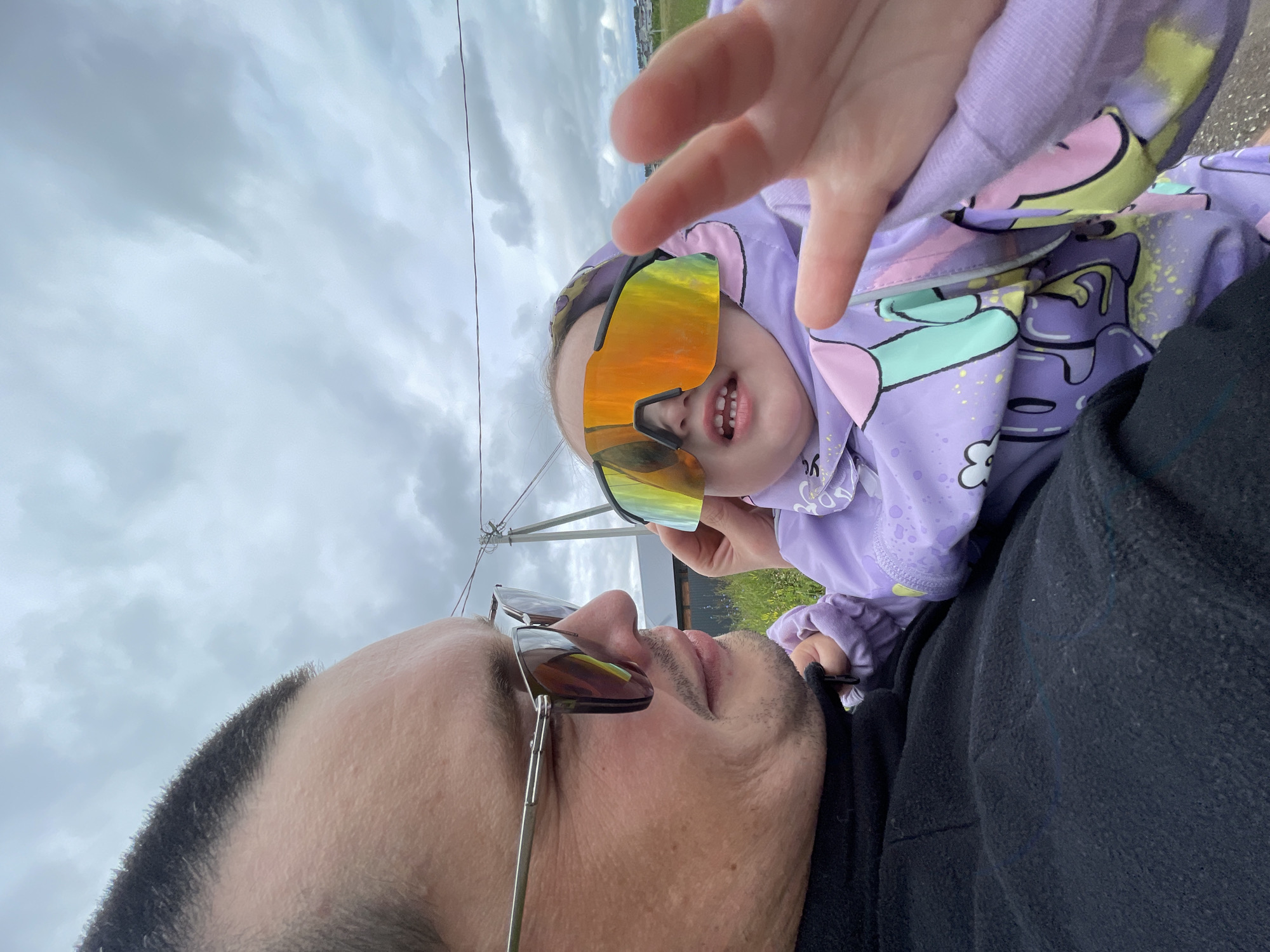 A low-angle close-up shot shows a man and a young child outdoors under a cloudy sky. The man, on the left, is wearing sunglasses with dark lenses and a black jacket. His face is turned upwards and slightly to the right, with a stubbled chin and mustache. The child, on the right, is wearing large, mirrored, multi-colored sunglasses that reflect the sky and surrounding scenery. The child is wearing a light purple hooded jacket with cartoonish graphics in pastel colors. The child's hand is reaching towards the camera, with fingers spread. The background consists of a gray, overcast sky and a hint of green grass and possibly some distant buildings, suggesting an outdoor, possibly rural, setting in Podshivalovo, Russia. The image has a casual, selfie-like feel, capturing a playful moment between the man and child.