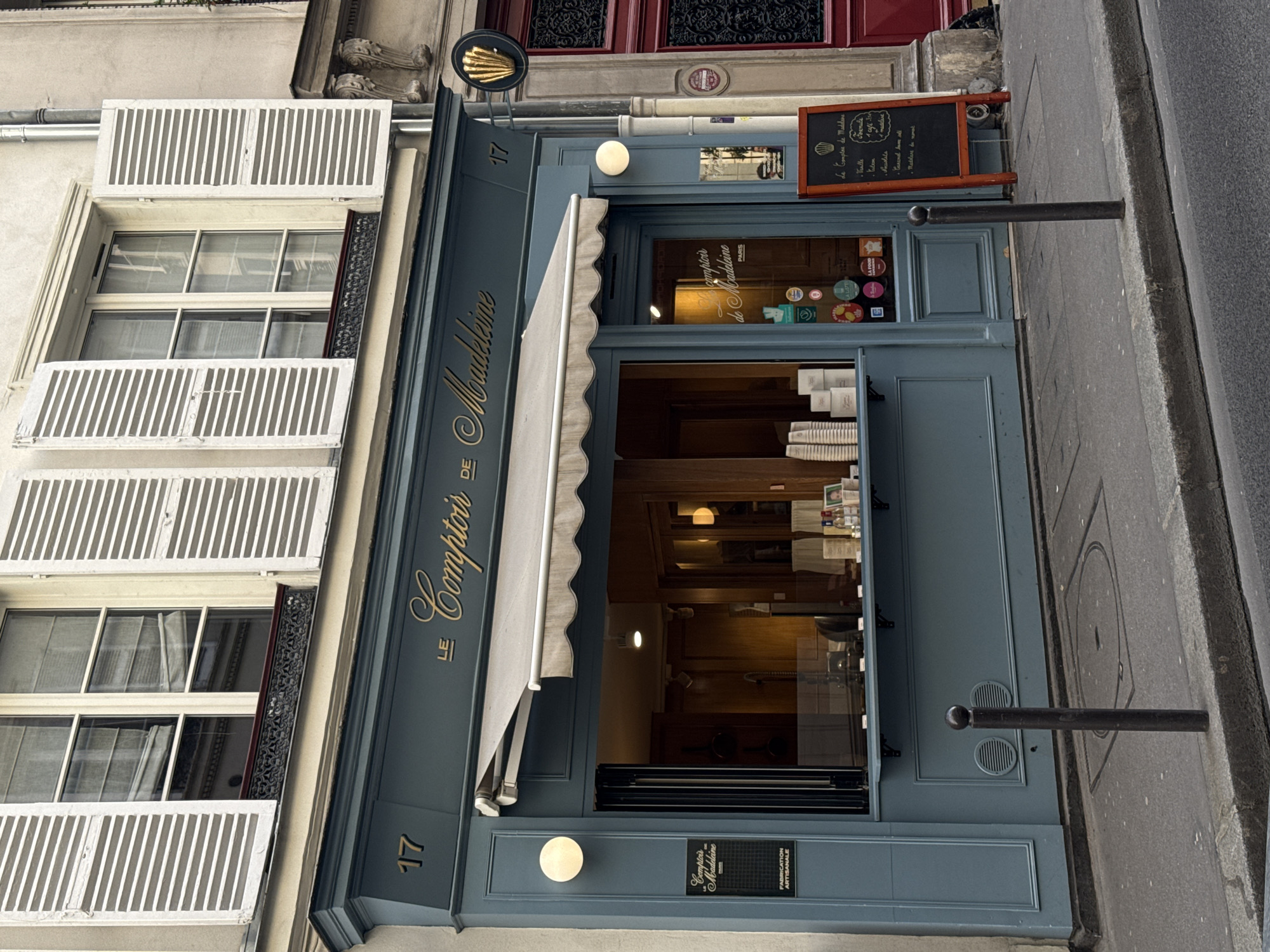 The image displays the facade of a charming shop, likely a patisserie or cafe, situated in a building on a city street, characteristic of Paris, France. The shopfront is painted in a distinctive dusty blue-grey color, with a white retractable awning extended over the entrance.
The shop's name, "LE Comptoir DE Madeleine," is prominently written in elegant gold script on the blue fascia above the awning. The number "17" is visible twice on the building's facade, indicating its address. Another sign on the lower right of the shop window also reads "Comptoir de Madeleine PARIS" and "FABRICATION ARTISANALE," suggesting artisanal production.
Through the large glass windows of the shopfront, the warm interior lighting reveals a cozy space with wooden finishes. Stacks of disposable cups are visible on a counter inside. On the right side of the entrance, an A-frame chalkboard menu displays items like "Le Comptoir de Madeleine," "1 café," and various flavors or items such as "Vanille," "Foenule," "Canard ham ali," and "Madeleine de nomad," reinforcing the establishment's specialty. Above the main entrance, a small golden sign shaped like a madeleine shell is affixed. Numerous circular stickers, possibly awards or ratings, are visible on the shop window.
Above the shop, the upper floors of the building feature residential windows with classic white louvered shutters, some open and some partially obscuring the windows. The building itself appears to be made of light-colored stone or stucco. The street level shows a grey sidewalk and a black metal bollard standing near the curb on the right. No people are visible, and the scene appears to be set during daylight hours on a clear day.