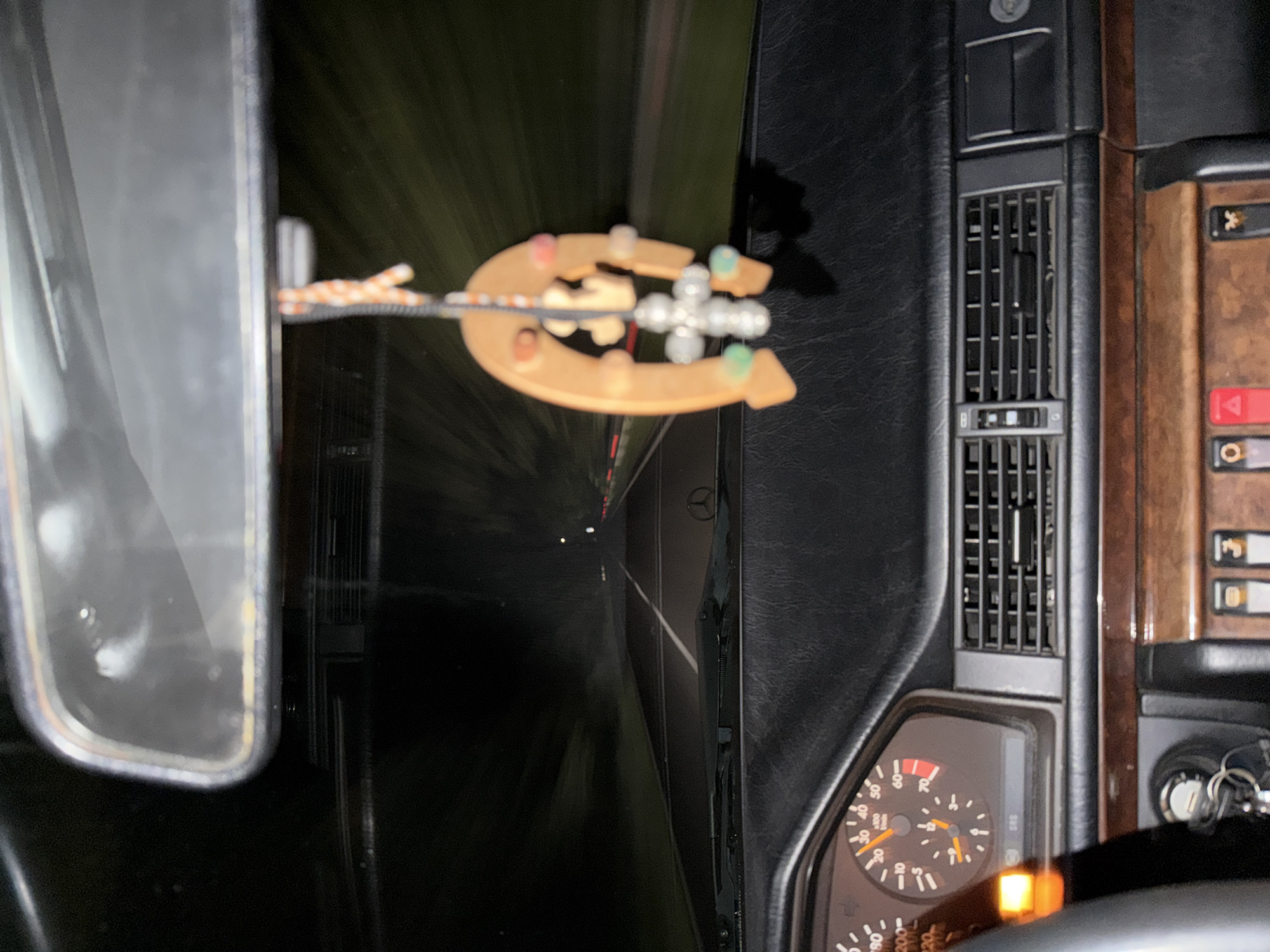 The image is taken from inside a car, possibly moving at night. The interior features a leather-clad dashboard, an air vent, and a wooden panel with switches. A speedometer is visible, showing the car is in motion. A rear-view mirror reflects the road. A horseshoe-shaped ornament hangs from the mirror. The road ahead is blurred, indicating movement. There is no clear indication of the location being Borovtsi, Bulgaria.