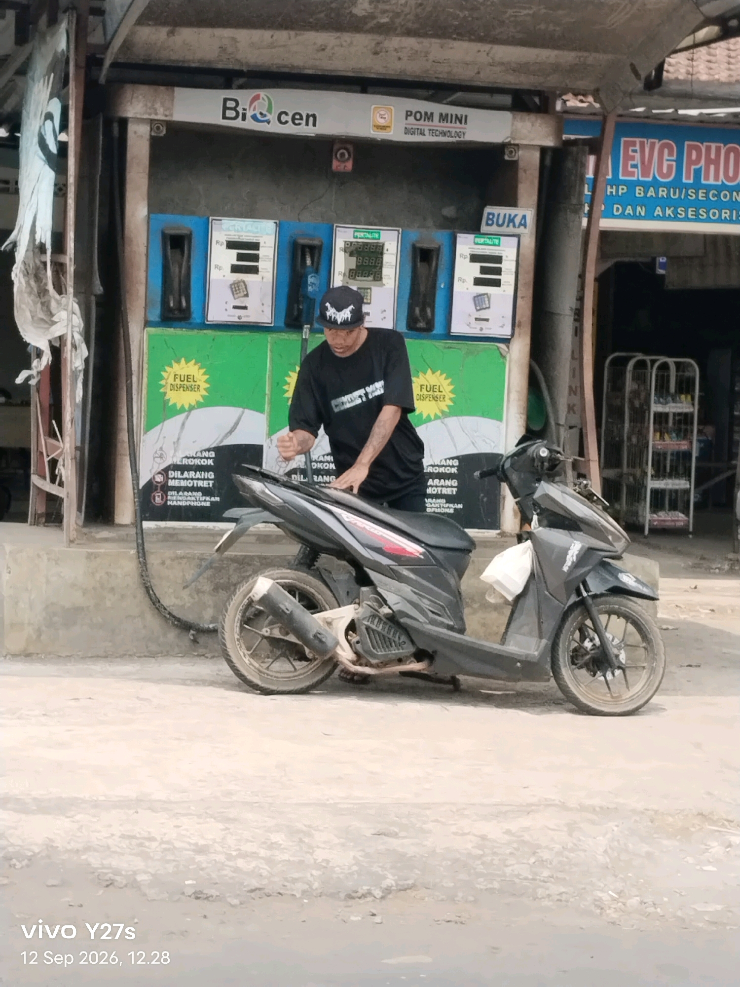 A person is standing next to a motorcycle at a gas station. The gas station has three pumps and signs that say "Bi-Cen," "POM MINI DIGITAL TECHNOLOGY," and "BUKA." The motorcycle is black and has a white bag on the front. The person is wearing a black t-shirt and a black baseball cap. They appear to be interacting with the motorcycle. The environment is a street in Trimulyo, Indonesia, with a concrete ground. The time and date stamped on the image indicate it was taken on September 12, 2026, at 12:28 PM.