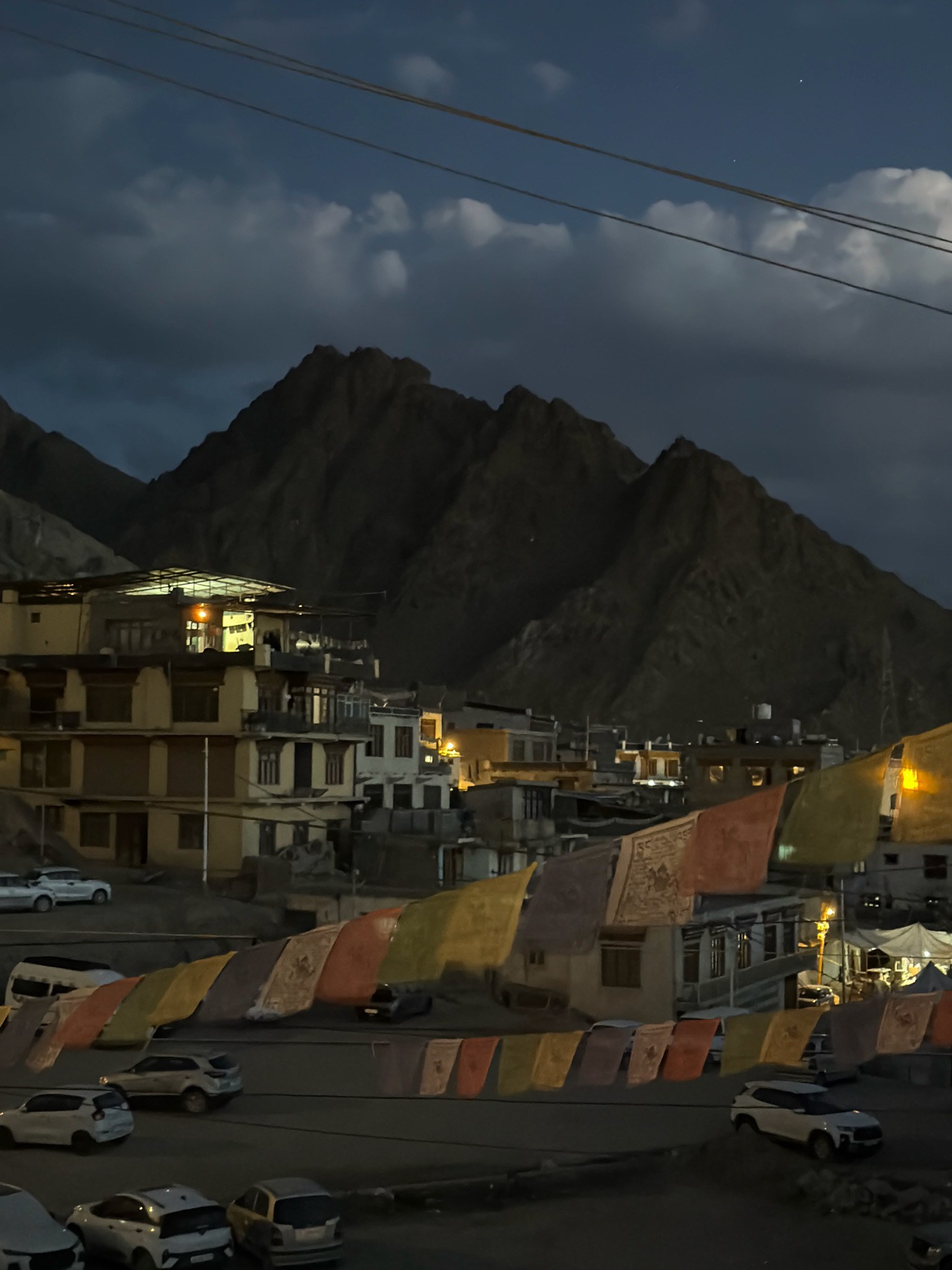 The image depicts a twilight scene in Leh, India, characterized by a cluster of buildings nestled against a backdrop of imposing mountains. Several vehicles, including cars and vans, are parked or driving on a road that runs in front of the buildings. Colorful prayer flags, a common sight in the region, are strung across the foreground, fluttering gently. The sky above is dark and cloudy, with a few faint stars visible, indicating either early dawn or late dusk. The buildings themselves appear to be a mix of residential and commercial structures, with lights visible in some of the windows, suggesting ongoing activity. The overall atmosphere is calm and serene, with the natural grandeur of the mountains contrasting with the urban elements of the town.