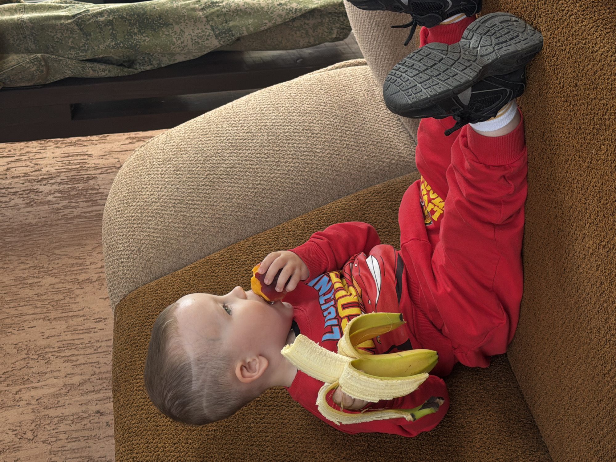 This image captures a young child lying on a textured brown sofa, with their feet propped up on a lighter beige sofa or cushion. The child is dressed in a vibrant red long-sleeved shirt and matching red pants. A graphic on the shirt, partially visible, appears to depict characters or branding associated with Disney's "Cars" movie, possibly Lightning McQueen.
The child is actively eating, holding a peach or nectarine to their mouth. Resting on their lap or chest is a peeled banana, split into two halves. The child's legs are raised, revealing black sneakers with distinct textured soles, worn over white socks with yellow stripes.
In the background, a wooden surface or floor is visible, along with a piece of camouflage fabric, suggesting an indoor, casual home setting, possibly in Chaikovsky, Russia. The textures of the sofas, the wooden surface, and the camouflage material are prominent. The child appears focused on their snack, creating a scene of relaxed and simple activity. No specific time of day or weather is discernible.
