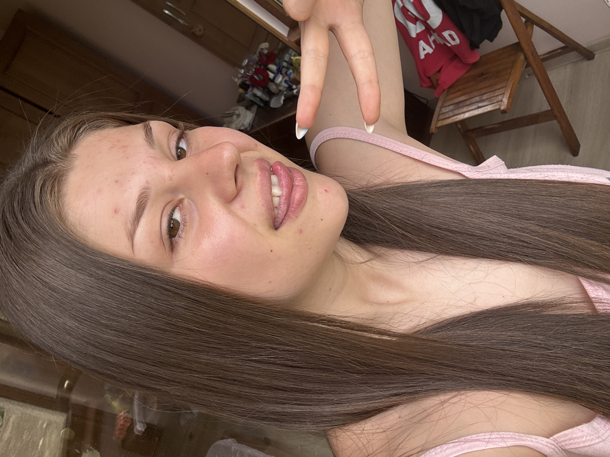 A close-up, sideways, full-body shot of a young person with long, dark brown hair. The person is wearing a light pink tank top. Their face is turned towards the camera, and they are making a peace sign with their right hand. The person has light-colored skin with some blemishes on their cheeks and forehead. Their lips are slightly parted, revealing their teeth. The background is out of focus, but it appears to be an indoor setting with a wooden chair and some clothing items hanging on a wall. The lighting suggests it is daytime, possibly indoors. The overall mood seems casual and playful. There is some visible text on the red clothing item in the background, appearing to be "AAD".