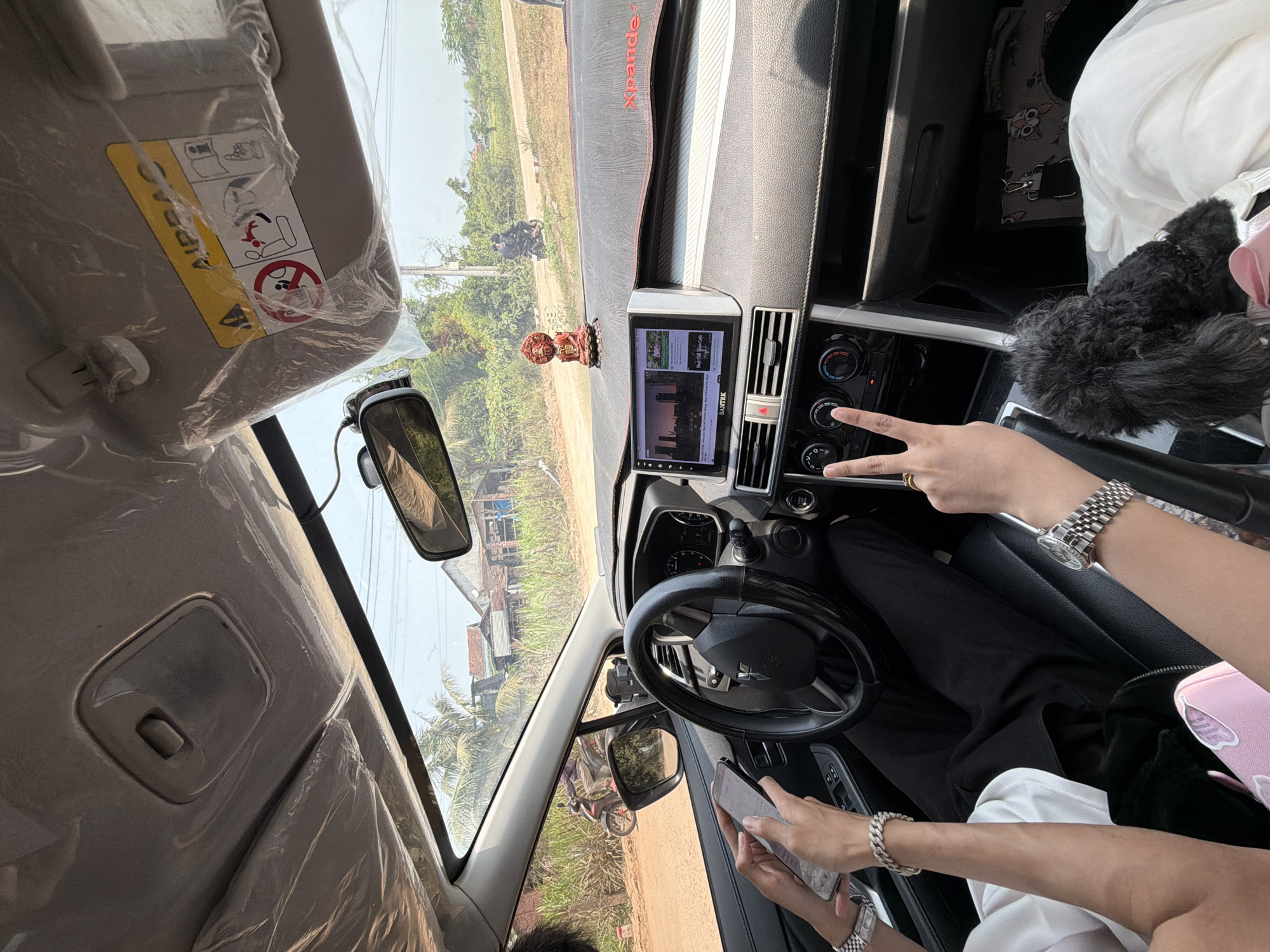 The image captures the interior of a car, looking out towards a rural or semi-rural landscape on a sunny day in Bình An, Vietnam. The car's interior appears relatively new, with plastic protective coverings still on the sun visor and parts of the roof lining.
On the dashboard, a multimedia screen displaying the brand "SANTEK" shows what looks like video content or a navigation screen, with visible Vietnamese text including "Thiết Kế Cảnh Quan." A small, reddish-brown decorative figurine, possibly a Buddha, sits centrally on the dashboard, which is covered by a mat bearing the text "Xpander." The steering wheel, partially visible, features a Mitsubishi logo. Below the multimedia screen are the car's air conditioning and audio controls.
At least two people are in the car. The hands of the person in the driver's seat are visible, one resting on the steering wheel and the other holding a smartphone. This person wears a silver watch and a matching bracelet. Near the central console, another hand, likely belonging to a passenger, makes a "peace" sign, adorned with a ring. Another wrist with a watch is also visible on the passenger side. A black fluffy object, possibly a small pet or a bag, and a white bag are on the passenger seat.
Outside, the view through the windshield and side mirror reveals a dirt road or path, lush green vegetation, distant trees, and simple structures or houses. A motorcycle with a person on it is visible outside. The sun visor clearly displays an "AIRBAG" warning label along with pictograms advising on passenger safety, particularly regarding children. The scene suggests a journey or a stop in a rural Vietnamese setting.