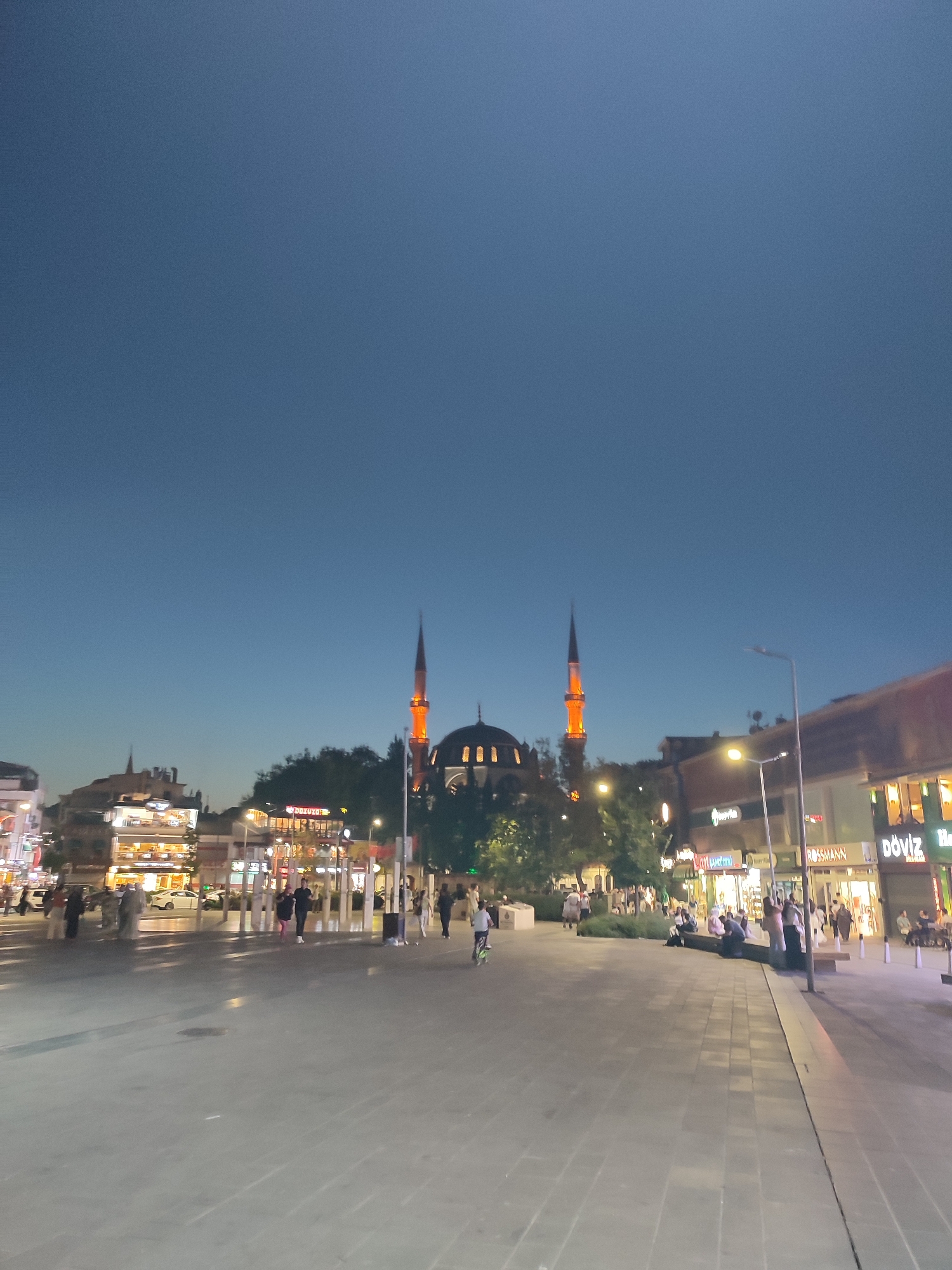 This is a nighttime, eye-level, medium-shot photograph of a wide, paved plaza in Üsküdar, Turkey. The plaza is lined with businesses and features a prominent mosque with illuminated minarets and dome. Several trees and streetlights are also visible. People are walking, standing, and sitting in the plaza. Some are interacting with each other, while others appear to be engaged in individual activities. The sky is a dark blue, indicating it is evening or night. The overall atmosphere is lively and urban.