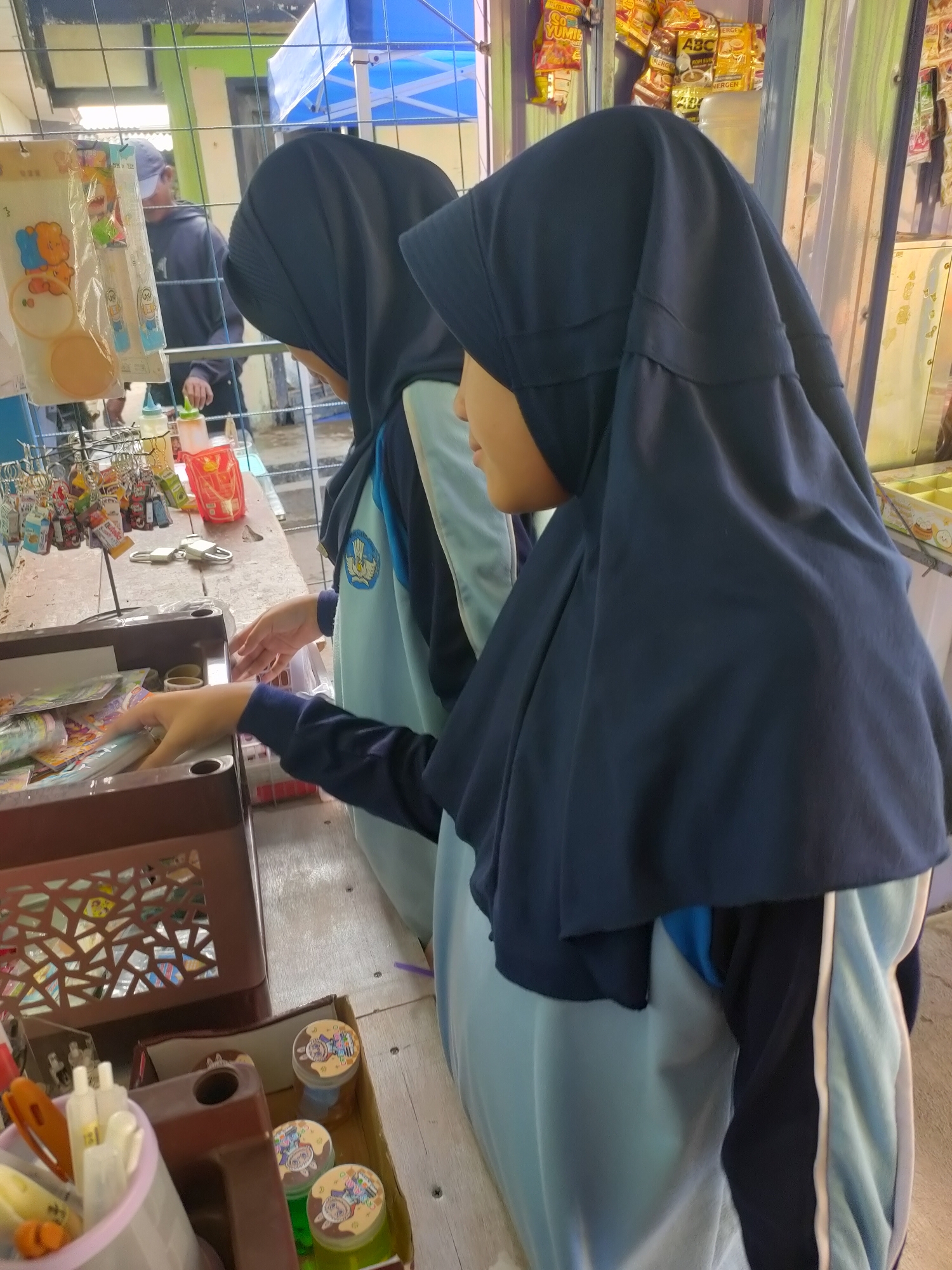 Two young Indonesian girls, wearing dark blue hijabs and light blue school uniforms with a school emblem, are standing at a stall in what appears to be an outdoor market or school fair in Subang, Indonesia. The girl in the foreground is reaching into a brown plastic container filled with various small items, possibly stationery or trinkets. The girl behind her is also looking at the items on the stall. The stall itself is cluttered with small consumer goods, including keychains, bottles of sauces or condiments, and packaged snacks displayed on shelves and in containers. In the background, a man wearing a dark hoodie and a cap is visible, and there are various colorful banners and a blue tent structure. The ground is made of concrete or pavement. The overall scene suggests a casual commercial setting, possibly during a school event.