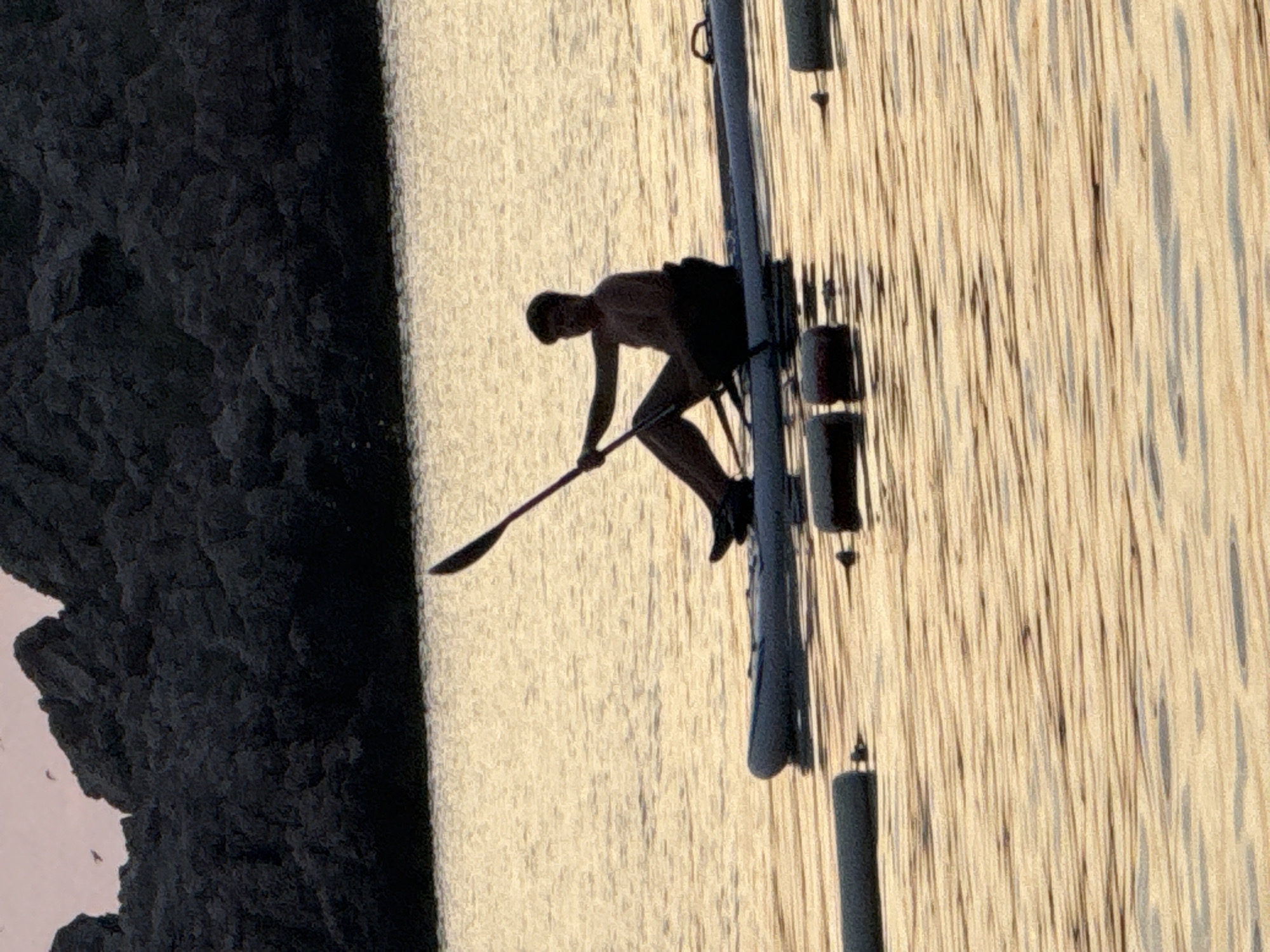 In the city of Pržno, Montenegro, a young person is silhouetted against the golden light of the setting sun as they paddle a stand-up paddleboard on calm water. The person, positioned in the center of the frame, is wearing dark shorts and is shirtless, holding a paddle with both hands. Their body is angled slightly towards the left, and their knees are bent as they sit on the paddleboard. The paddleboard, a light gray inflatable model, floats on the shimmering water. In the foreground, several dark, cylindrical buoys are visible, tethered to the seabed, breaking the surface of the water. In the background, a dark, rocky shoreline rises dramatically, covered in dense vegetation. The sky above the shoreline is a soft pink, and a few small birds are faintly visible in flight. The overall impression is one of peaceful recreation at dusk.