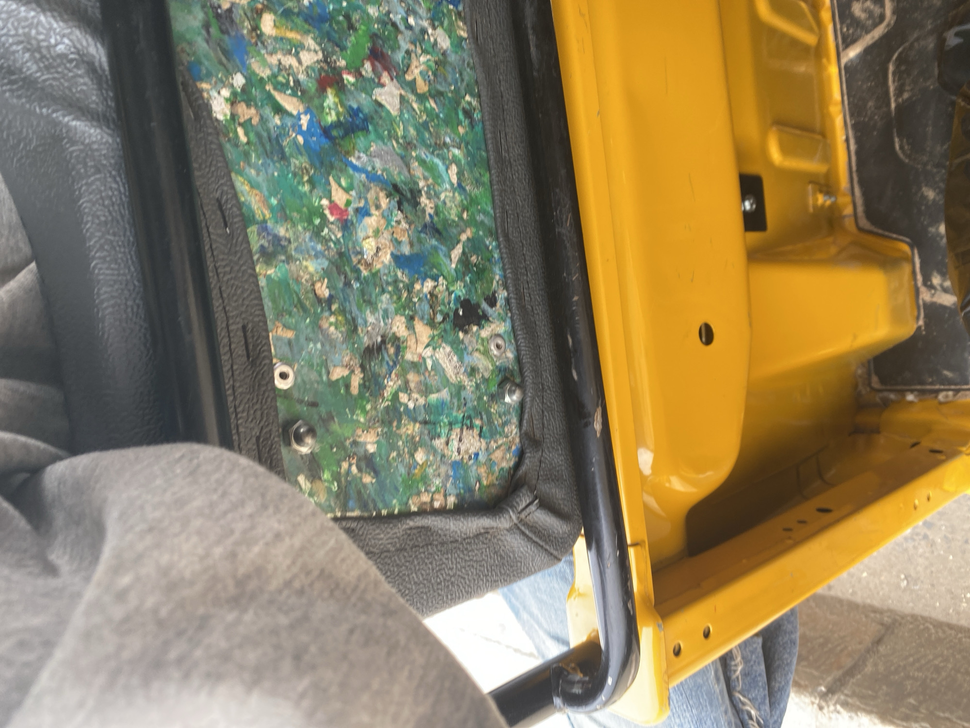 An overhead shot captures the interior of a bright yellow three-wheeled auto-rickshaw, likely taken in Jalingo, Nigeria. A person's legs, clad in blue jeans, are visible on the left, suggesting they are seated in the vehicle. The focus is on the back of the passenger seat, which is upholstered in black material and displays a unique, multicolored composite material underneath, appearing to be made of recycled fragments. The composite material is a mix of greens, blues, reds, and beige flecks, secured with visible metal grommets. A black metal bar, part of the auto-rickshaw's frame, runs horizontally across the top of the seat. The yellow body of the auto-rickshaw is visible below the seat, with some fasteners and structural elements of the vehicle also in view. The ground beneath the auto-rickshaw is a mixture of sand and debris.
