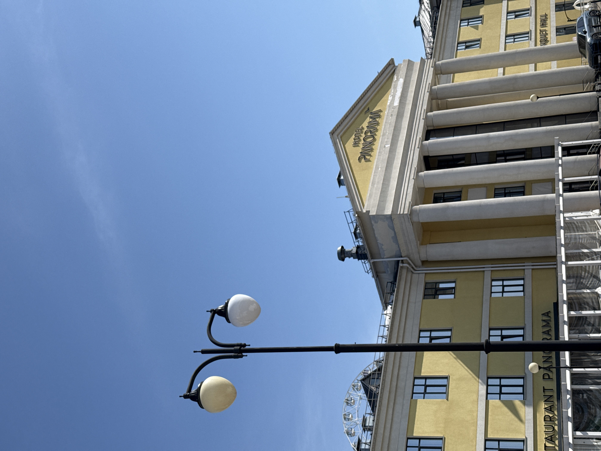 The image shows an exterior view of the Panorama Hotel and Restaurant in Izhevsk, Russia, under a clear blue sky. A black street lamp with two white globular lights is in the foreground. The building is light yellow with a geometric, art deco style and prominent horizontal features. Hotel PANORAMA is inscribed at the top, and RESTAURANT PANORAMA is vertically aligned on the side of the building. A Ferris wheel is partially visible behind the building. A black car is parked on the right edge of the frame.