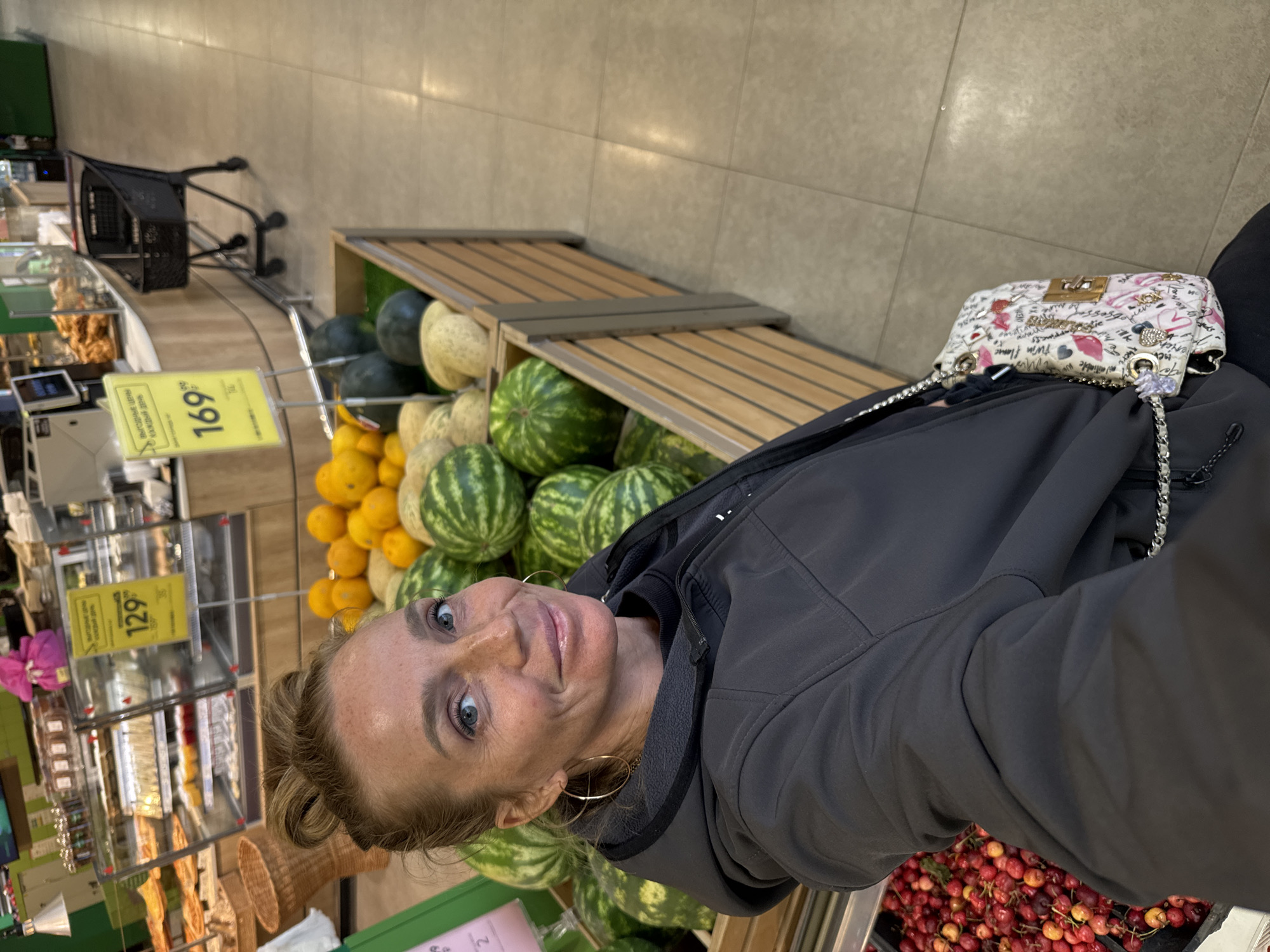 A medium-shot, high-angle selfie shows a fair-skinned woman with blonde hair pulled up in a bun, looking at the camera. She is wearing a dark gray hooded jacket and a small white purse with a chain strap adorned with pink drawings. The woman is in a supermarket in Mistolovo, Russia. Behind her, there are bins filled with watermelons and melons, as well as a display of oranges. Price tags indicating 129 and 169 are visible. In the background, there are bakery displays and a shopping cart. To the left, a bin of red cherries is partially visible.