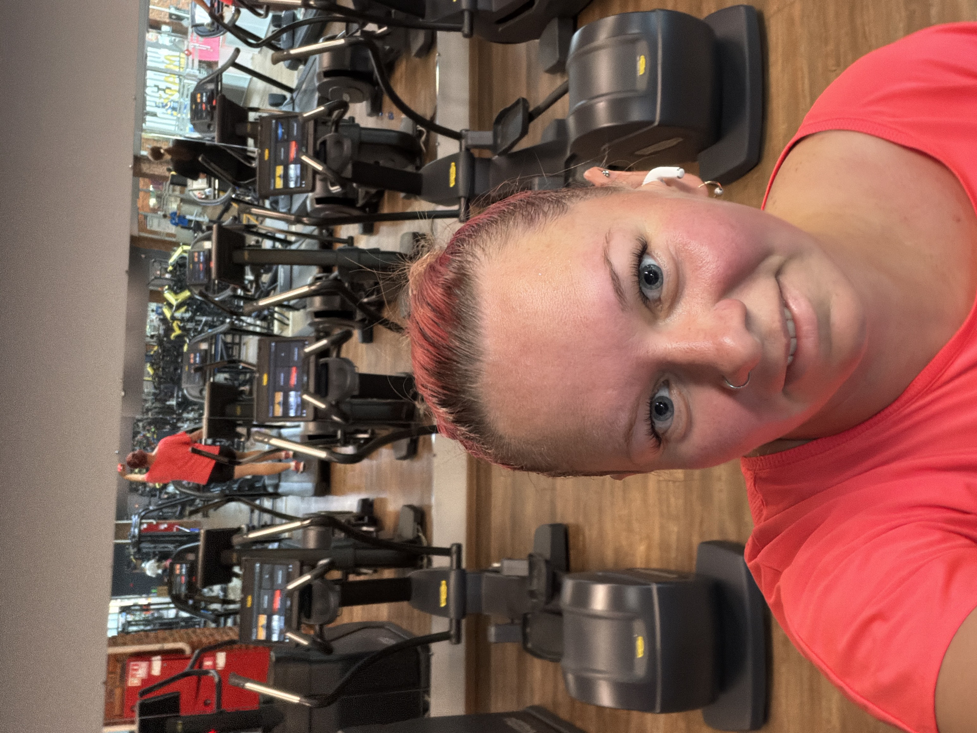 A woman with pink-tinted hair and blue eyes takes a selfie in a gym. She is wearing a bright coral t-shirt and has a nose ring and earrings. In the background, a row of elliptical machines is visible. A reflection in the mirror behind the elliptical machines shows other gym equipment and people exercising. The gym has wooden flooring and large mirrors on the walls. The location is a gym in the city of Chaikovsky, Russia.