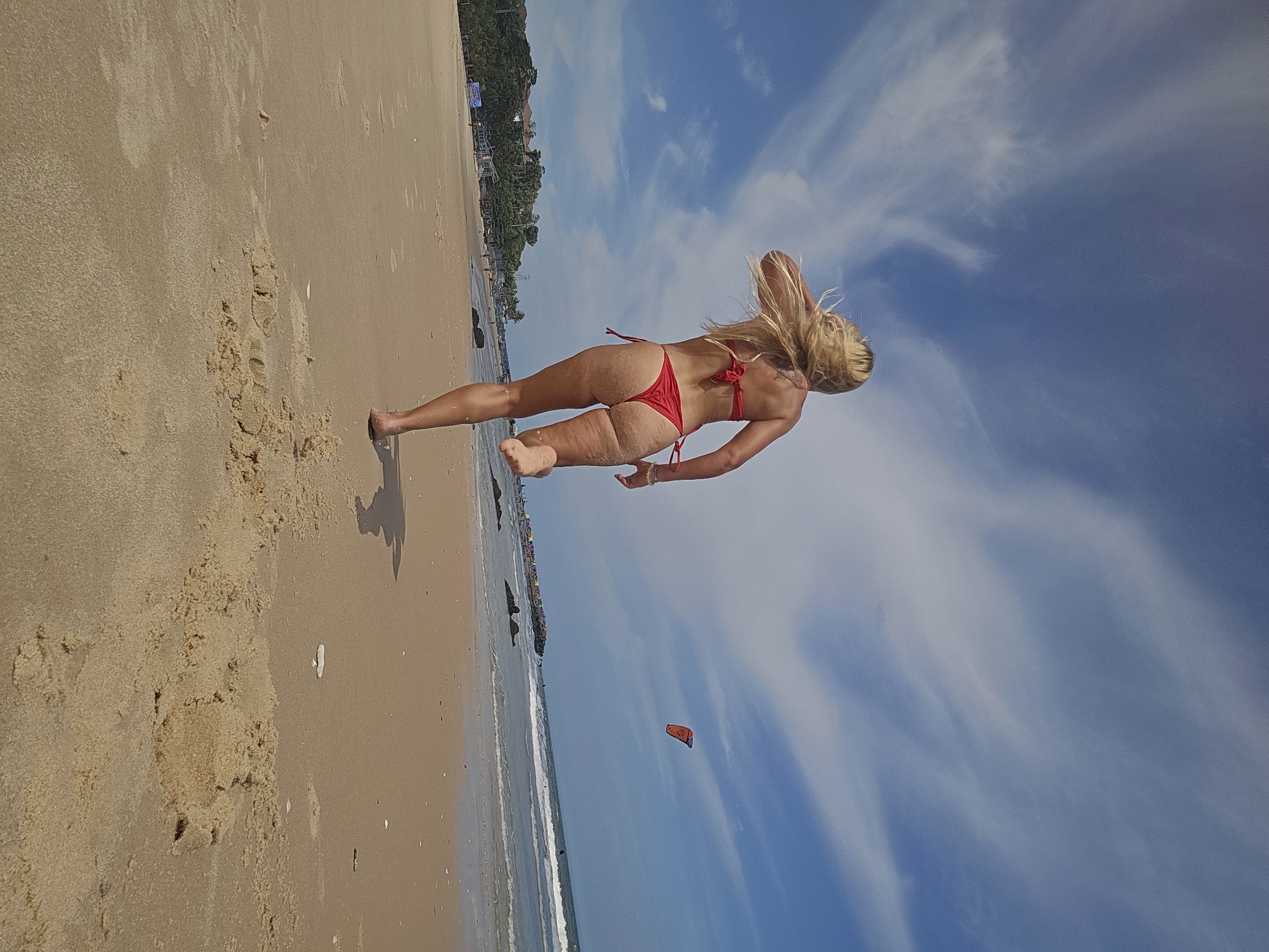 An image captures a woman in a red bikini running along a sandy beach. The scene is set against a backdrop of a bright blue sky with wispy clouds. In the distance, the ocean waves are visible, and a kite surfer can be seen in the air. The beach appears to be a public space, with a line of umbrellas and some buildings visible along the coastline. The woman's hair is blonde and flows behind her as she runs, and her body is partially covered in sand. The overall impression is one of a sunny day at the beach.