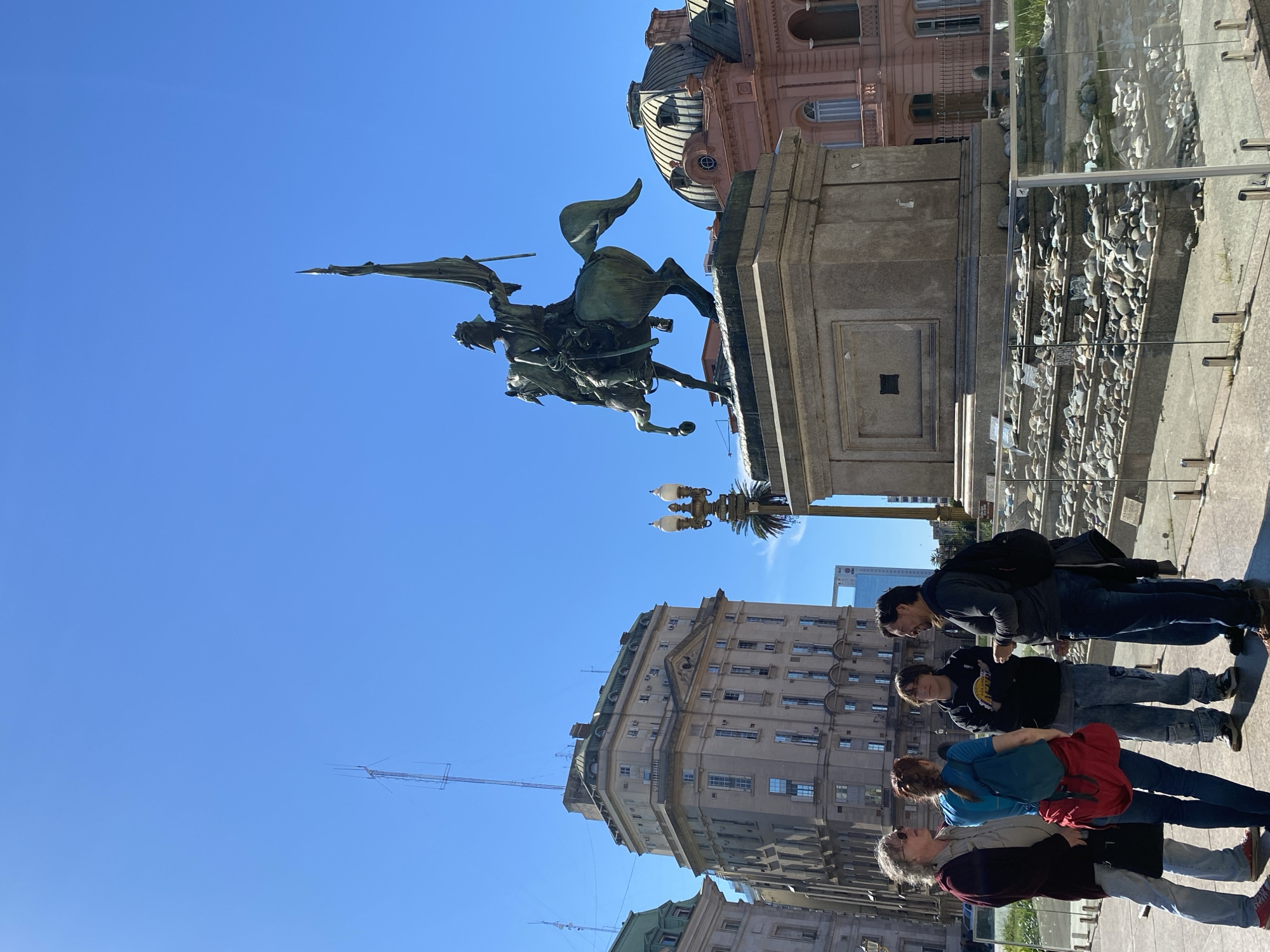 An outdoor scene in a bustling city square on a clear, sunny day. The dominant feature is a large equestrian statue of a historical figure, mounted on a tall stone pedestal, positioned on the right side of the frame. The statue depicts a person on horseback, holding a flag or standard, with intricate details. Behind the statue, a distinctive pink-hued building with domes and ornate architecture stands prominently against a bright blue sky, identifiable as the Casa Rosada (Presidential Palace).
In the foreground, to the left, a group of five individuals, appearing to be tourists or sightseers, are gathered. They are dressed in casual clothing and seem to be observing the statue and the surrounding architecture. Just beyond the statue's base, there is a low display area filled with numerous small stones and what appear to be commemorative objects, protected by a clear glass barrier. Further in the background, other city buildings, including a modern high-rise, contribute to the urban landscape. The bright sunlight casts clear shadows, indicating midday or early afternoon. The overall impression is one of a vibrant historical landmark, likely Plaza de Mayo in Buenos Aires, Argentina. No discernible text is legible on any signs or objects.