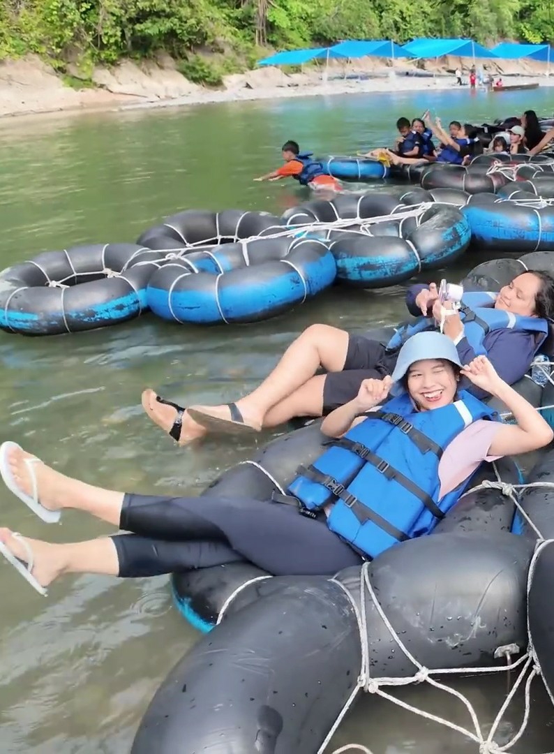 In the foreground, two women are relaxing on inflatable tubes in a river. The woman on the right is wearing a blue life vest and a dark blue jacket, and she is holding a cell phone. The woman on the left is wearing a blue life vest and a light pink shirt, and she has her hands up in the air. Both women are smiling and appear to be enjoying themselves. In the background, several other people are visible on inflatable tubes in the river. The river is surrounded by lush green trees and vegetation, and there are blue tents set up on the shore. The overall scene suggests a recreational activity in a natural setting.