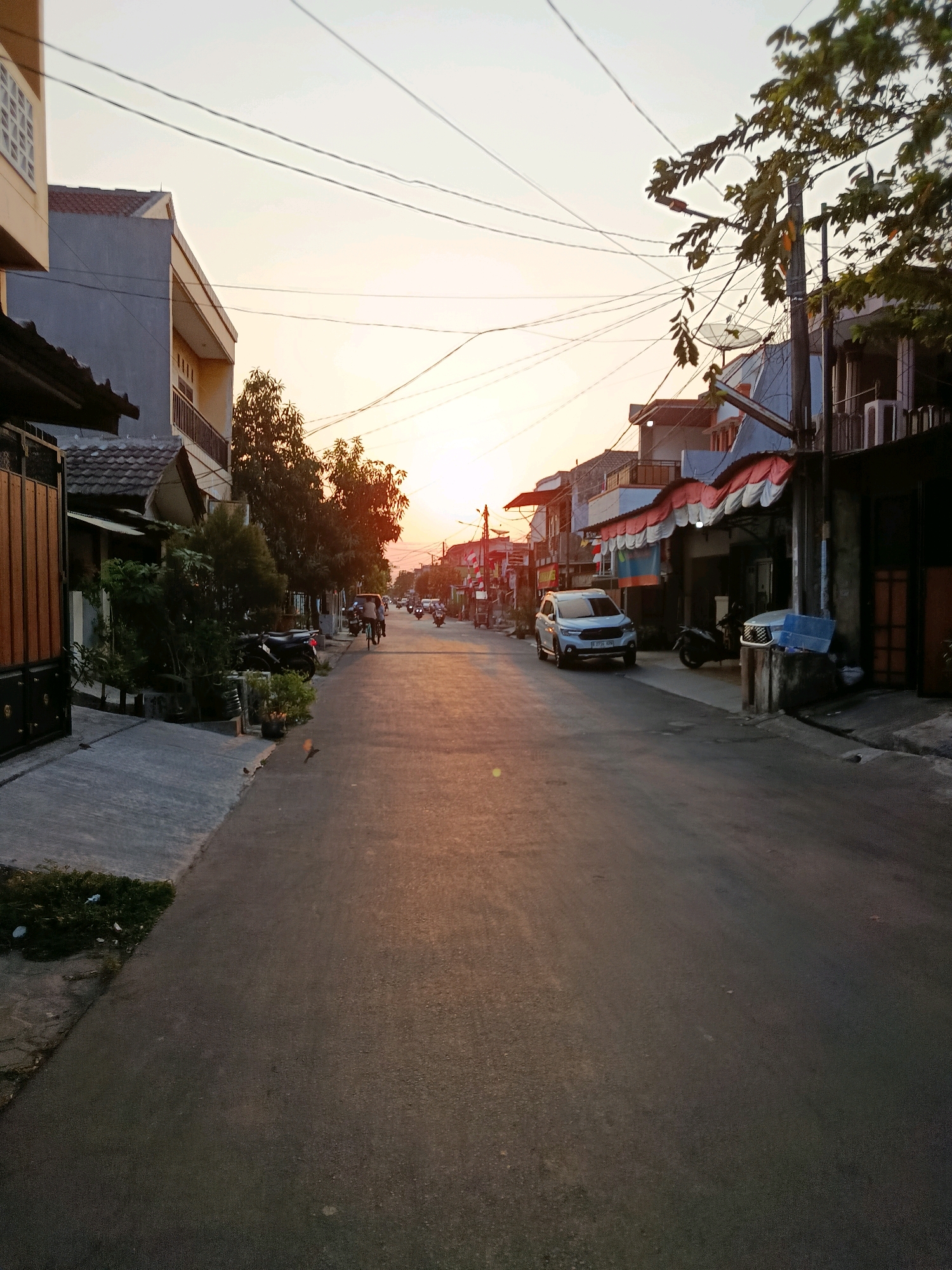 This image depicts a street scene in Bekasi, Indonesia, at dusk. The sun is setting, casting a warm glow on the asphalt road and the surrounding buildings. The street is lined with residential houses and shops, some with awnings and balconies. There are several vehicles on the road, including cars, motorcycles, and a bicycle. People are visible on the street, some riding vehicles and others walking. Overhead, a network of electrical wires crisscrosses the sky. The overall atmosphere is peaceful and quiet, suggesting a typical evening in a suburban area of Bekasi.