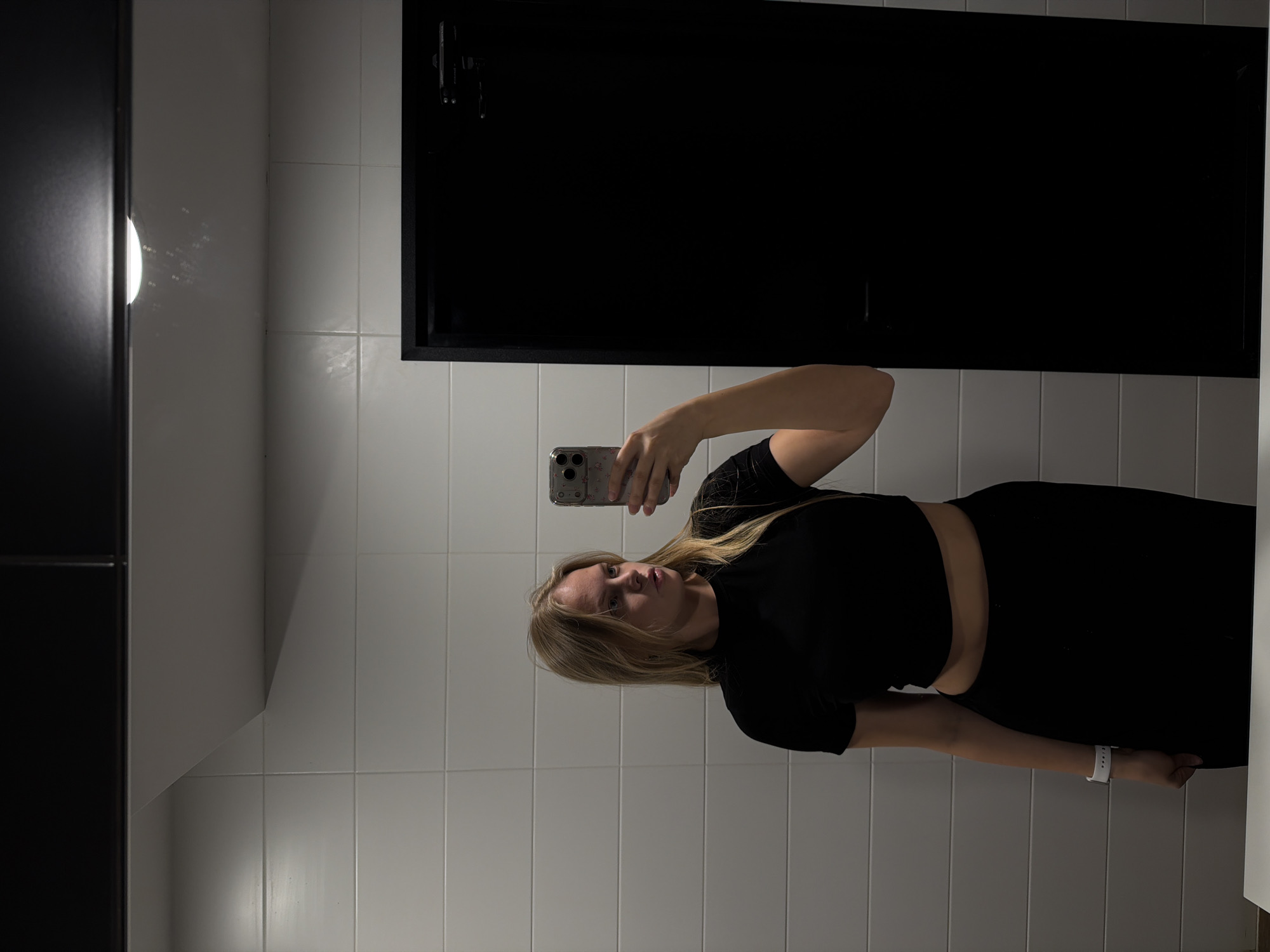 A young woman is depicted in a mirror selfie, holding a smartphone with a clear, patterned case in her right hand. She has long, straight blonde hair and a fair complexion, looking directly towards the camera with a neutral expression. She wears a black short-sleeved crop top and black bottoms, exposing a portion of her midriff. On her left wrist, a white smartwatch is visible, with faint text that appears to spell "friends" or a similar word.
The setting is an indoor space with white, square-tiled walls. A prominent dark rectangular fixture, possibly a large mirror frame or a dark reflective panel, occupies the upper background. On the far left, a circular light fixture illuminates the wall, creating subtle reflections and shadows in the room. The lighting is artificial and moderately bright, suggesting an indoor environment like a public restroom or changing room. The image was captured in Izhevsk, Russia. The time of day and weather are not discernible from the indoor setting.