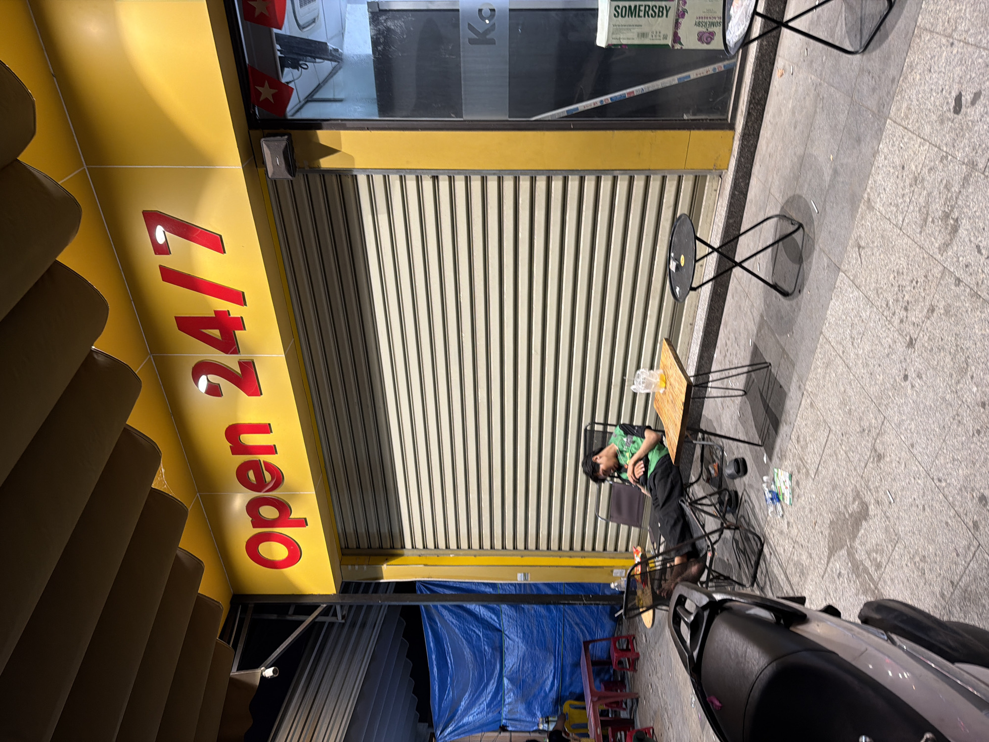 The photograph depicts a street scene in Đà Nẵng, Vietnam. A man is sitting at a small outdoor table, next to the closed roll-up door of a business marked "open 24/7". Two small tables sit next to him, and a motorcycle is parked nearby. A blue tarp stretches across the back of the building. Additional seating is visible beneath the tarp. The surrounding environment includes a paved sidewalk, and an adjacent building facade.