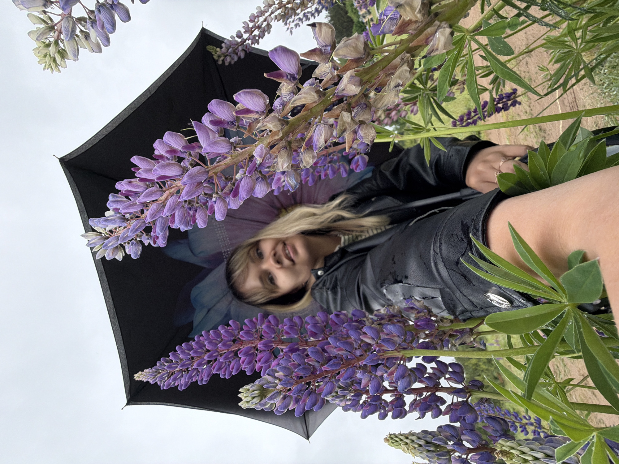 A woman is seen from a low angle, with her face framed by purple lupine flowers. She is smiling and holding a black umbrella with a flower design on the inside. She wears a black leather jacket. The sky is overcast. The location is near Chaikovsky, Russia, based on the presence of lupine flowers commonly found in the region.