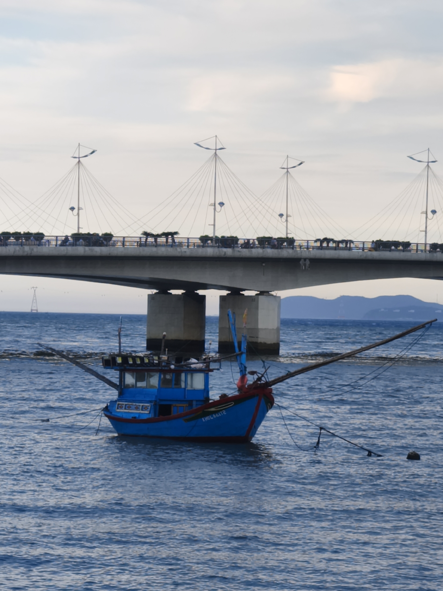 A vibrant blue fishing boat bobs on the choppy, deep blue water. The boat, with red trim and a cabin, has a long wooden outrigger extending to the right. Two poles rise from the deck, one with a blue flag or sail attached. Lights are visible on the cabin roof. Text is visible on the side of the boat: "KHÔI NẮNG 2".
In the background, a large concrete bridge spans the water, supported by substantial pillars. The bridge deck is lined with planters filled with green foliage. Cables from tall, slender poles extend to the bridge, creating a suspension structure. Several small figures, possibly people, are visible on the bridge. Further in the distance, a hazy mountain range meets the horizon under a pale, overcast sky. The overall scene suggests a coastal location in Vietnam, likely during daylight hours with cloudy weather.
