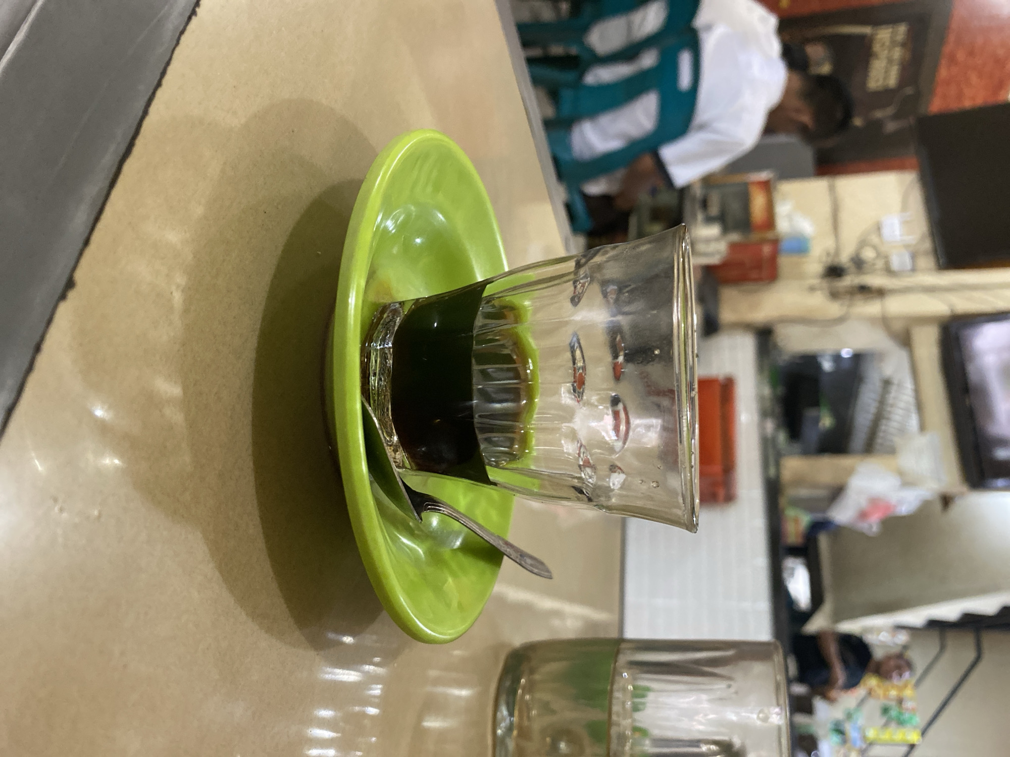 The image captures a close-up of a glass of dark liquid, likely coffee, placed on a bright green saucer. A small silver spoon rests on the saucer, partially tucked under the glass. The setting appears to be a casual eatery or cafe, with blurred figures and elements of a bustling environment visible in the background. To the left, a person wearing a white shirt is seated, and behind them, teal-colored chairs are lined up. In the background, there are shelves with various items, electrical wiring, and what looks like a television screen. To the right, another glass, partially filled with a lighter-colored liquid, is visible. The overall lighting suggests an indoor setting with ambient light sources. This scene is located in Lhokseumawe, Indonesia.