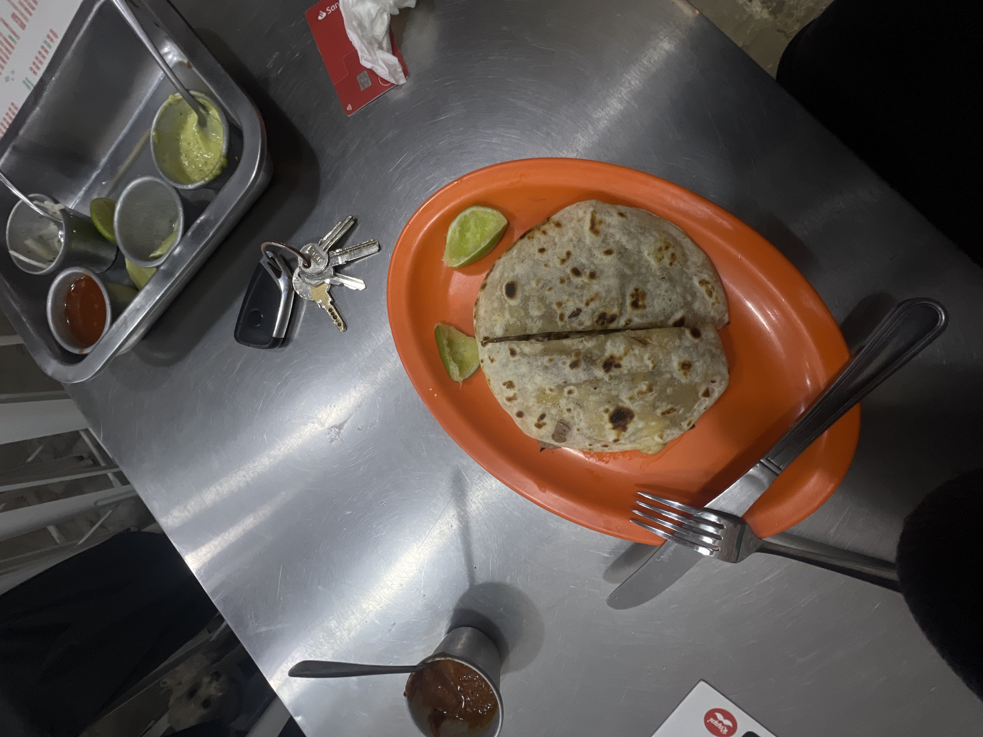 On a metallic tabletop, likely in a diner or food establishment, sits a partially eaten quesadilla on an orange plate, with lime wedges. A silverware set is beside it. There is a tray with multiple sauces in the corner. A set of keys are adjacent to the sauces. The image also shows a Santander card, and a small container of sauce with a spoon. The setting is likely Mexico City, Mexico, given the type of food and the presence of a Santander card, as Santander is a major bank in Mexico. The scene captures a casual meal setting.