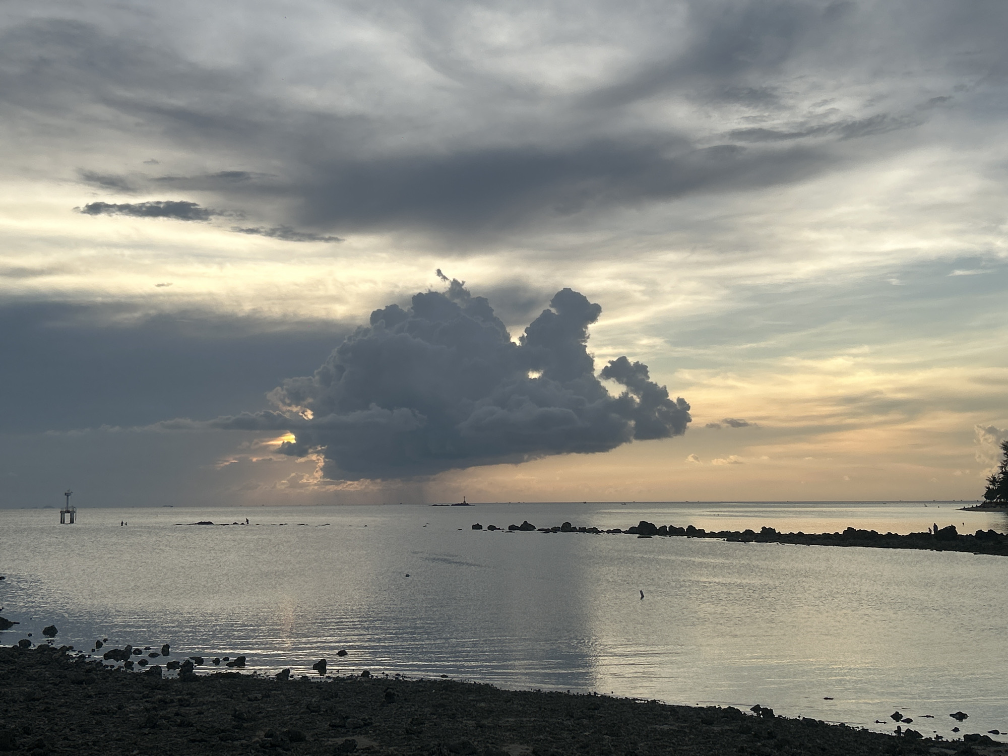 The image shows a coastal scene in Baan Sri Thanu, Thailand, under a cloudy sky. A large, dark cumulonimbus cloud dominates the center of the frame, partially obscuring the setting sun. The sea is calm, with a few scattered rocks and a navigational buoy visible. A low-lying, rocky shoreline is in the foreground. There are no people clearly visible.