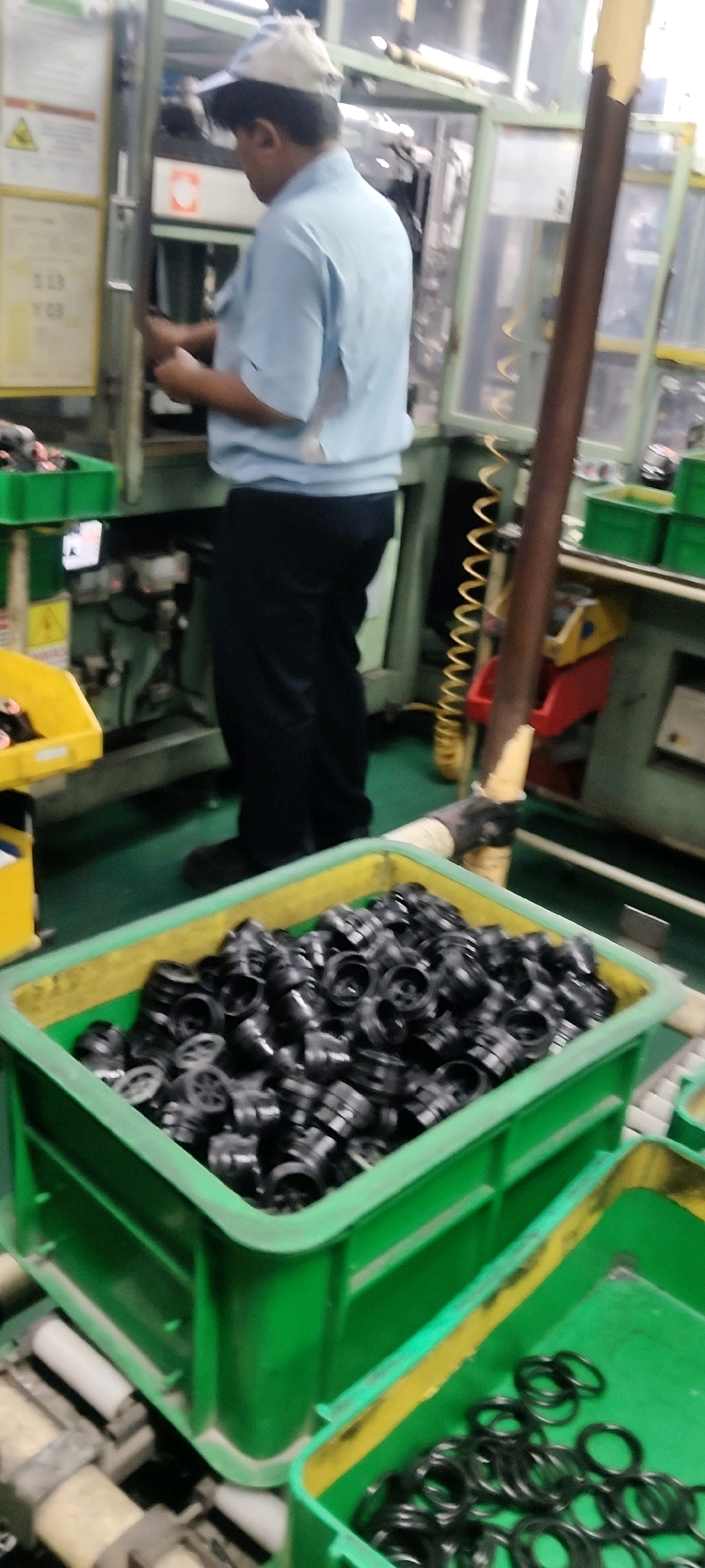 The image depicts a person wearing a light blue shirt and dark pants, with a light-colored cap, operating machinery in an industrial setting. The person appears to be standing and looking down towards the machinery, with their hands positioned near the controls. In the foreground, there are several green plastic bins containing black, circular components. One bin is partially filled with these components, while another, to the right, contains a scattering of similar-looking black rings. The background shows industrial equipment with various pipes and wires, suggesting a manufacturing or assembly environment. The overall scene is brightly lit, and the image is slightly blurred, indicating movement or a low-resolution capture. There is no visual information in the image that allows for the determination of a specific city or location like Mekarmukti, Indonesia.
