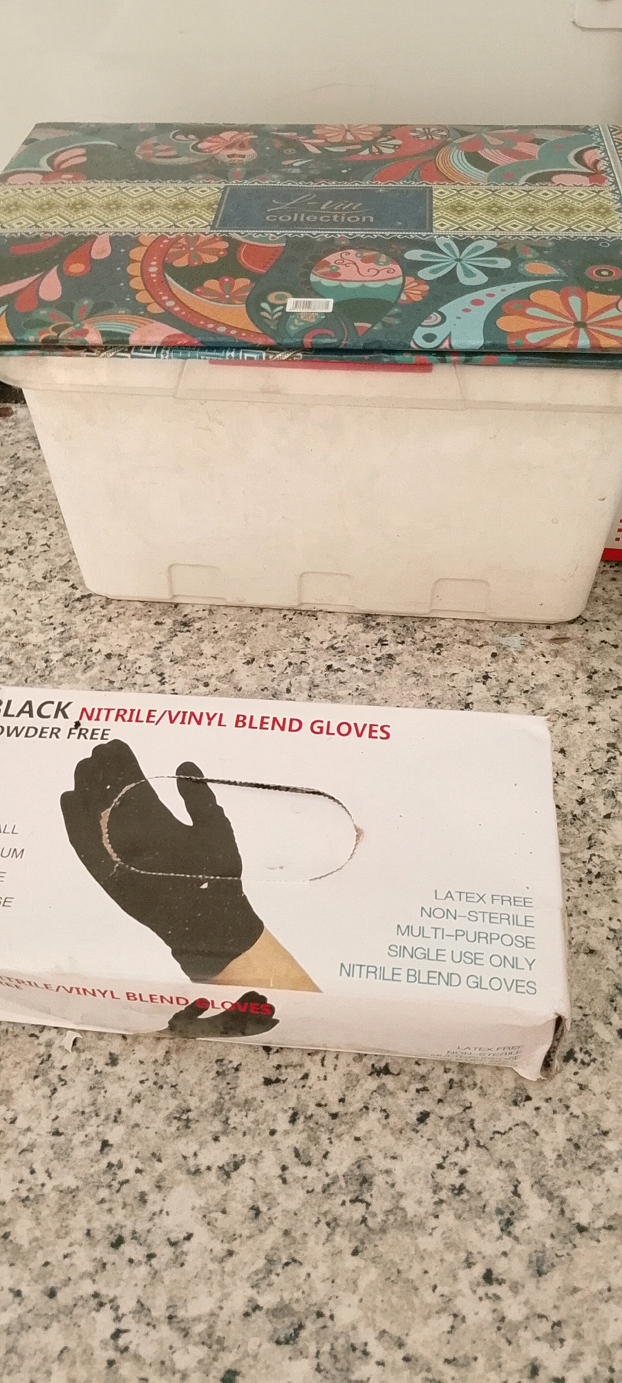 The image shows a close-up of a box of black nitrile/vinyl blend gloves on a speckled countertop. The box has text that reads "BLACK NITRILE/VINYL BLEND GLOVES" and "POWDER FREE". There is also an illustration of a gloved hand. To the right of the glove illustration, there is text that says "LATEX FREE", "NON-STERILE", "MULTI-PURPOSE", "SINGLE USE ONLY", and "NITRILE BLEND GLOVES". In the background, there is a white storage container and the lid of a colorful patterned box. The lighting is natural and appears to be from overhead. The image is taken from a slightly elevated angle, looking down at the box. The environment appears to be indoors. There are no people visible in the image. The specific location in the City of Medan, Indonesia cannot be determined from the provided image.