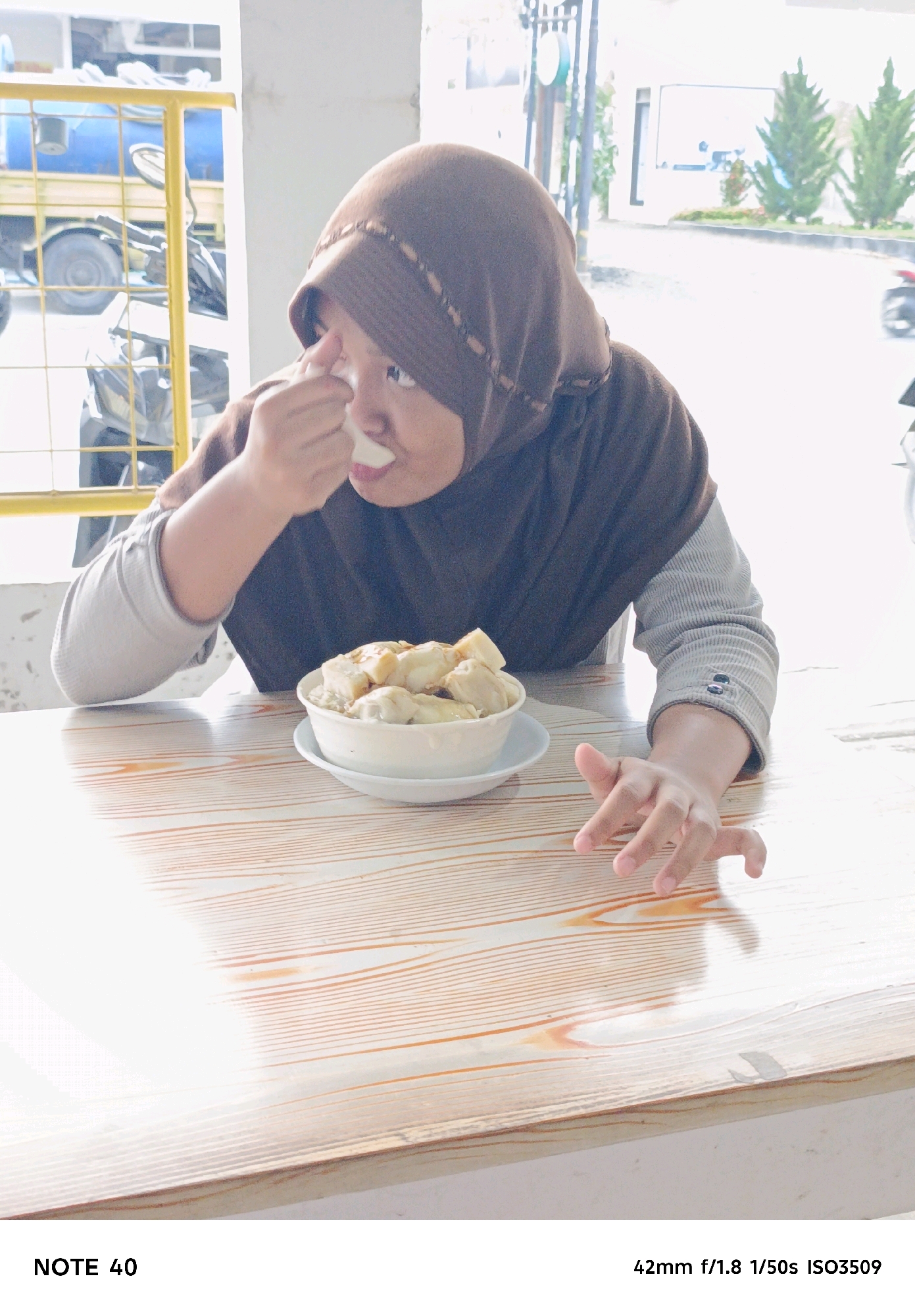 A person wearing a brown hijab and a gray long-sleeved shirt is eating from a bowl of food on a wooden table. The person is holding a spoon in their right hand and bringing it to their mouth. Their left arm is resting on the table. In the background, there is a yellow metal structure, a blue truck, and some trees. The scene appears to be outdoors.