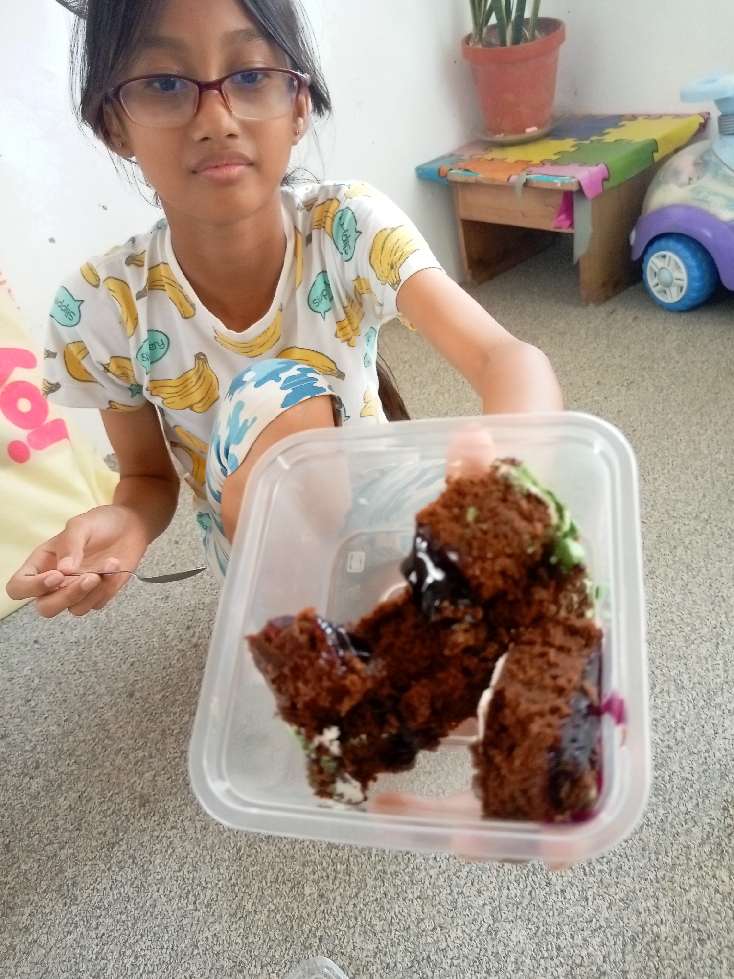 An outdoor portrait shows a young Filipina girl with glasses, dark hair pulled back, and a white t-shirt with a banana print, holding a clear plastic container with a slice of chocolate cake. The girl is on her knees on a gray carpeted surface, with her right arm extended forward, presenting the cake. The cake appears to have green frosting and dark, syrupy filling. In the background, there is a small wooden bench with a colorful puzzle mat on top, a potted plant, and a toy car. The scene appears to be in a residential setting in Batam, Indonesia, given the commonality of such home environments in the area. The lighting suggests it is daytime.