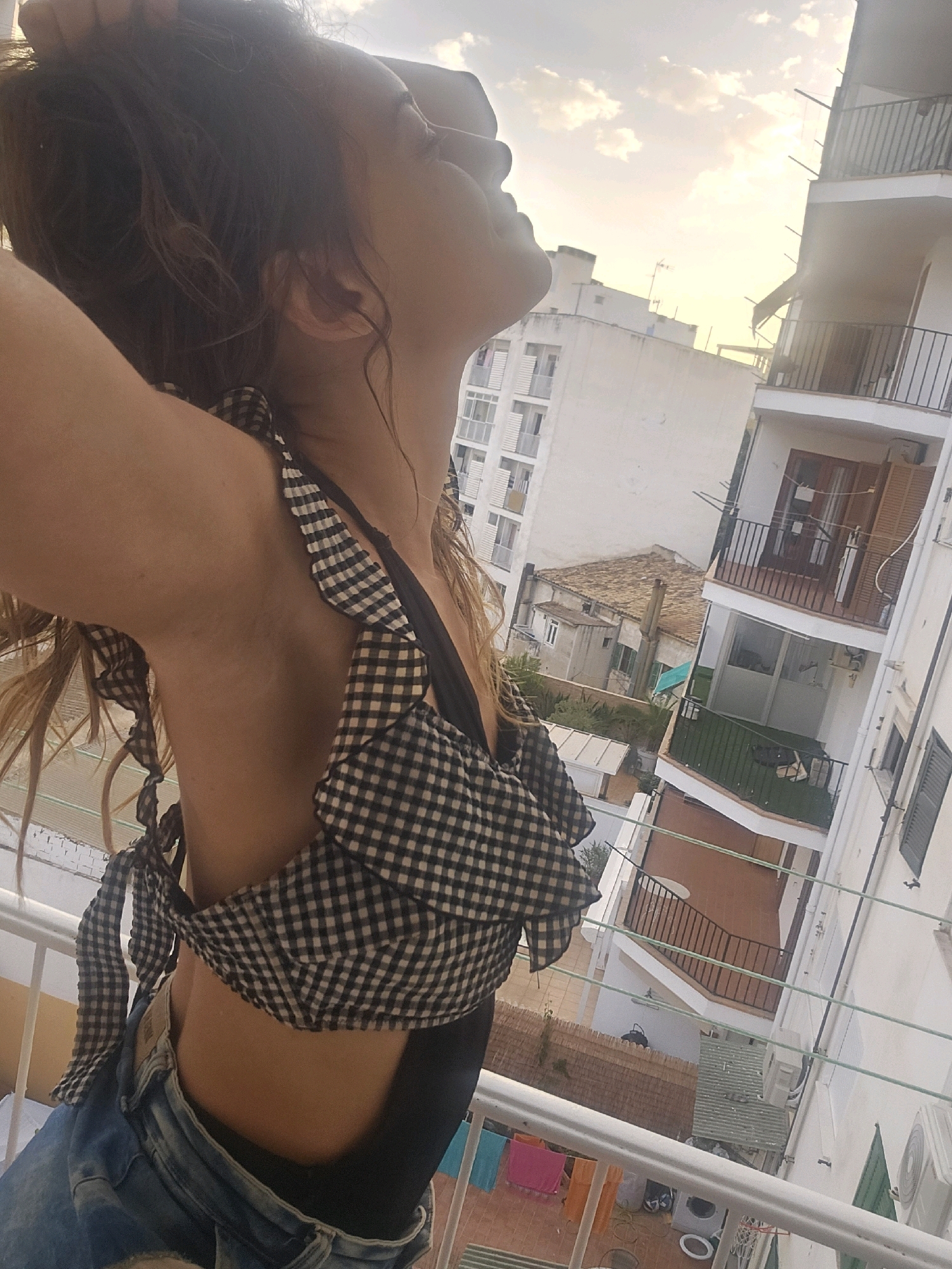 A woman is pictured from the chest up, looking upwards and to her right, with her left arm raised and hand touching her head. She is wearing a black and white gingham-patterned top with ruffled trim and blue denim jeans. She stands on a balcony overlooking a city street with white buildings and balconies. The sky is cloudy with the sun setting. The photograph appears to have been taken during golden hour in Palma de Mallorca, Spain.