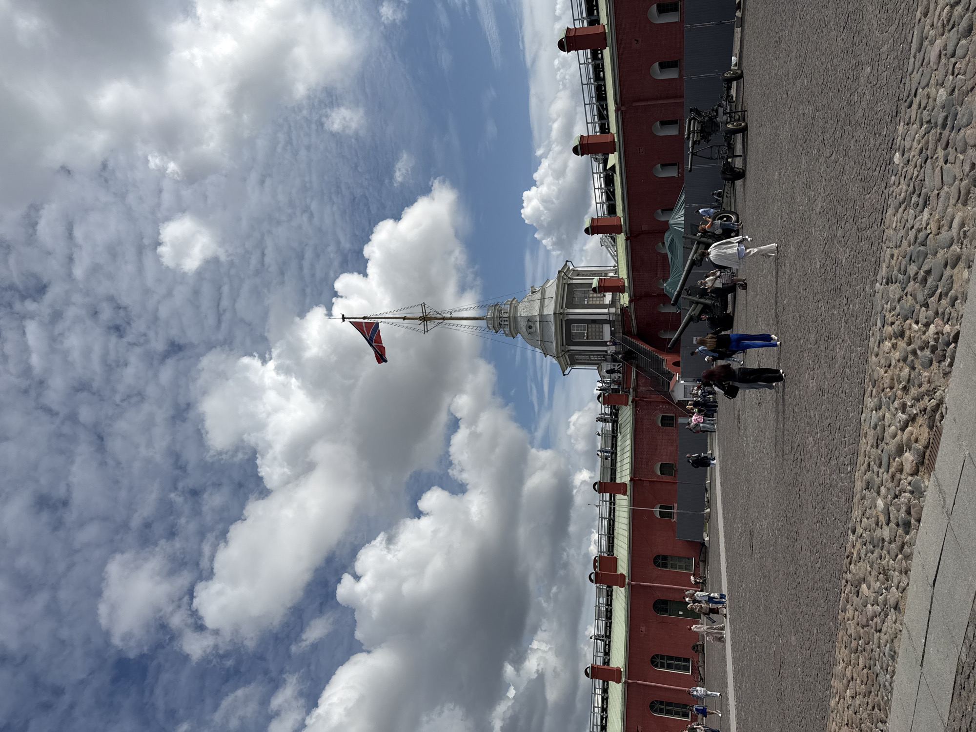 The image captures a wide-angle view of the Peter and Paul Fortress in Saint Petersburg, Russia. The foreground is paved with cobblestones and shows several people walking across the courtyard. In the middle ground, a red brick fortress wall with arched windows stretches across the frame, topped with a green roof and decorative red chimneys. A central tower, octagonal and grey with a white spire, rises above the fortress, topped by a flagpole with a distinct flag, likely the Russian naval ensign. Two historical cannons are positioned in the courtyard to the right of the central tower, one larger and the other smaller. The sky is a mix of blue patches and dramatic, fluffy white clouds. Several people are visible on the ramparts of the fortress, observing the scene. The overall impression is of a historically significant site on a bright, cloudy day.