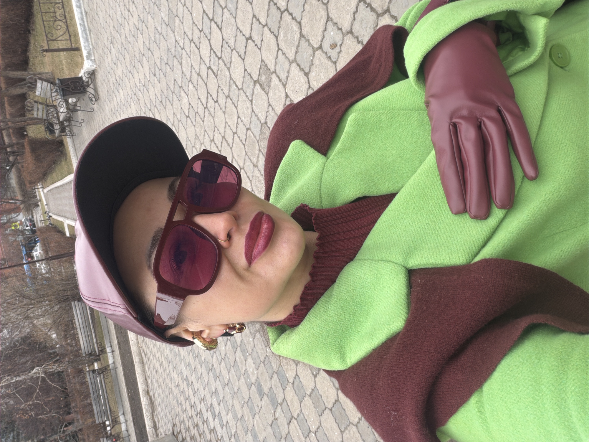 The image shows a woman wearing a dark burgundy leather cap, matching sunglasses and gloves, a burgundy turtleneck sweater, and a lime green coat with burgundy trim. She is outdoors on a paved area, possibly a sidewalk, with a patterned stone design. In the background, there are benches, trees, and buildings, suggesting an urban park setting. Based on visual cues like the style of the benches and the general appearance of the environment, the location could be Krasnoyarsk, Russia, as suggested. The woman's face is in focus, displaying matching lipstick and accessories.