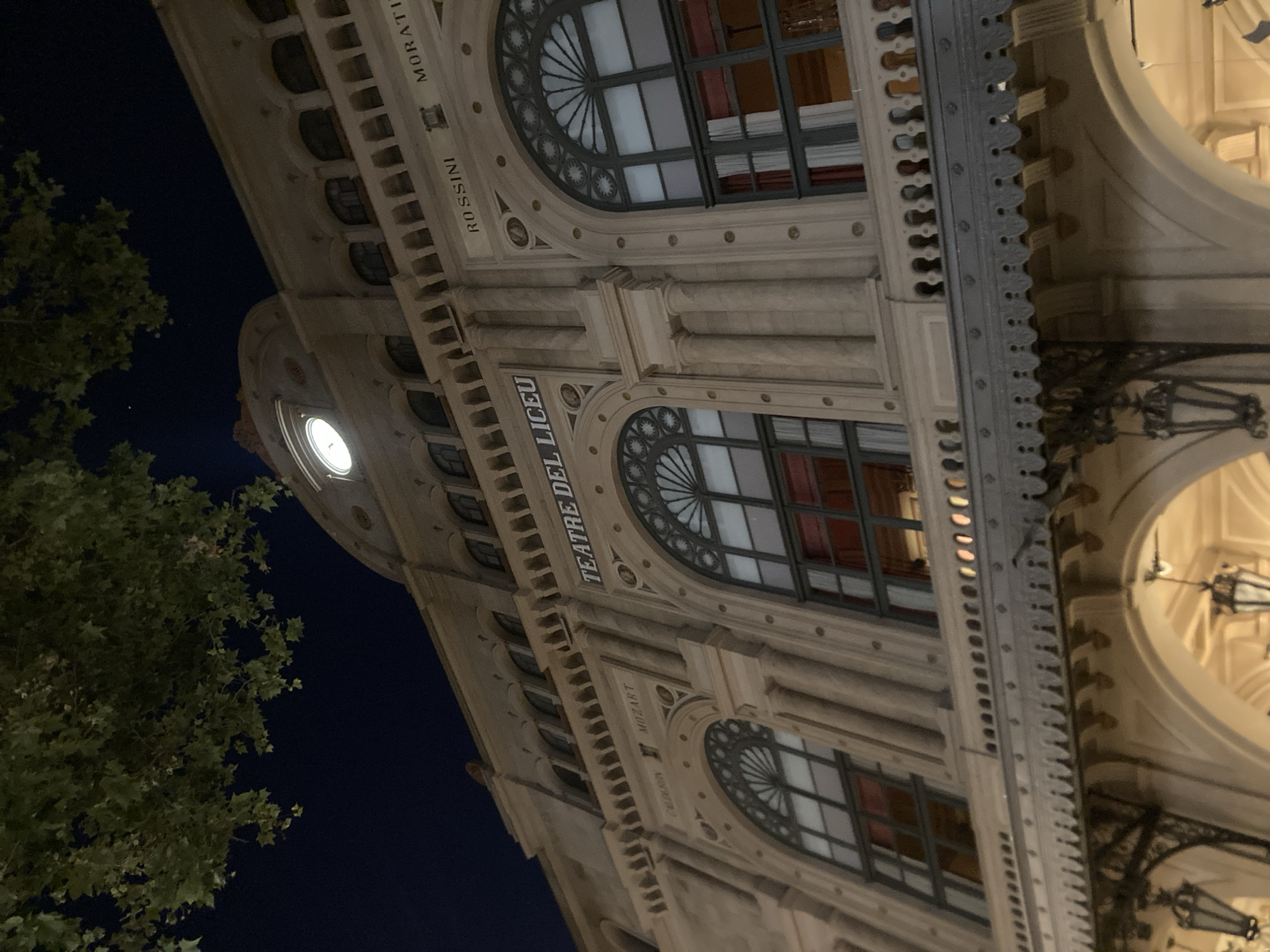 The image shows a close-up, tilted view of the exterior of a grand building at night. The building appears to be made of stone and features classical architectural elements, including arched windows with decorative grilles, ornate cornices, and columns. A prominent clock face is visible on the left side of the building, illuminated from within. Above some of the windows, text is inscribed. Visible text includes "TEATRE DEL," "LYCEU," "ROSSINI," and "MORATI." The scene is illuminated by artificial lights from within the building and from street lamps, casting shadows and highlighting the architectural details. A large, leafy tree occupies the left side of the frame, obscuring some of the building's facade and reaching into the dark night sky. The overall impression is one of historical grandeur and evening elegance. The location is identified as Barcelona, Spain, which, along with the text, suggests this is likely the Liceu opera house.