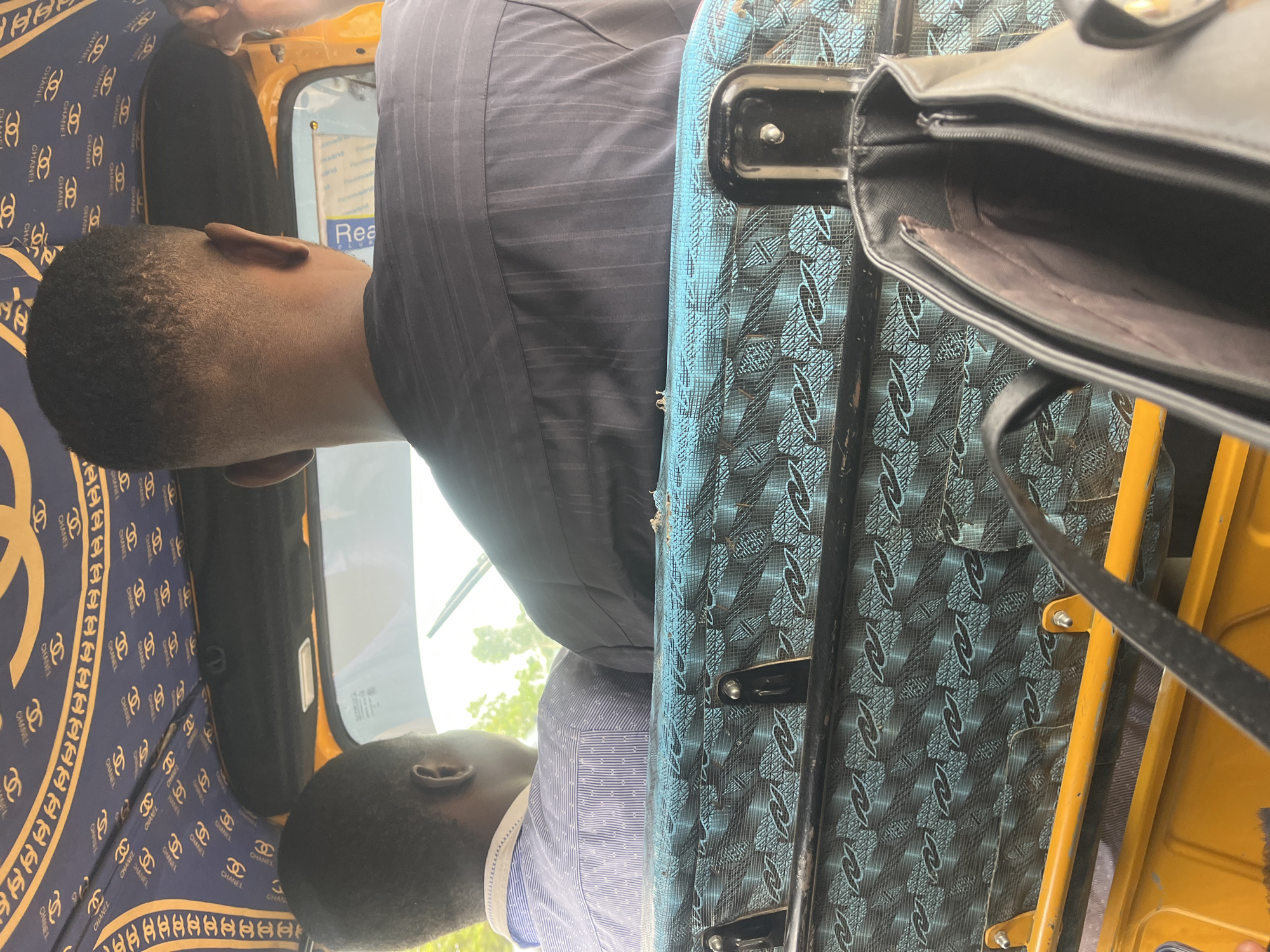 The image shows the interior of a commercial vehicle, likely a tuk-tuk, in the city of Karum, Nigeria. Two individuals are seated in the vehicle, viewed from behind. The person in the foreground is wearing a dark, pinstriped shirt, while the person in the middle ground wears a blue patterned shirt. The interior of the vehicle is decorated with a blue fabric featuring a repeating pattern of gold "CC" logos, resembling a Chanel design. The seats are covered in a teal fabric with a black and silver interlocking pattern. A black handbag with a silver zipper is visible in the foreground, resting on a yellow surface, possibly part of the vehicle's frame. The overall lighting suggests it is daytime, and the background shows a blurry view of greenery, indicating an outdoor urban or semi-urban environment.