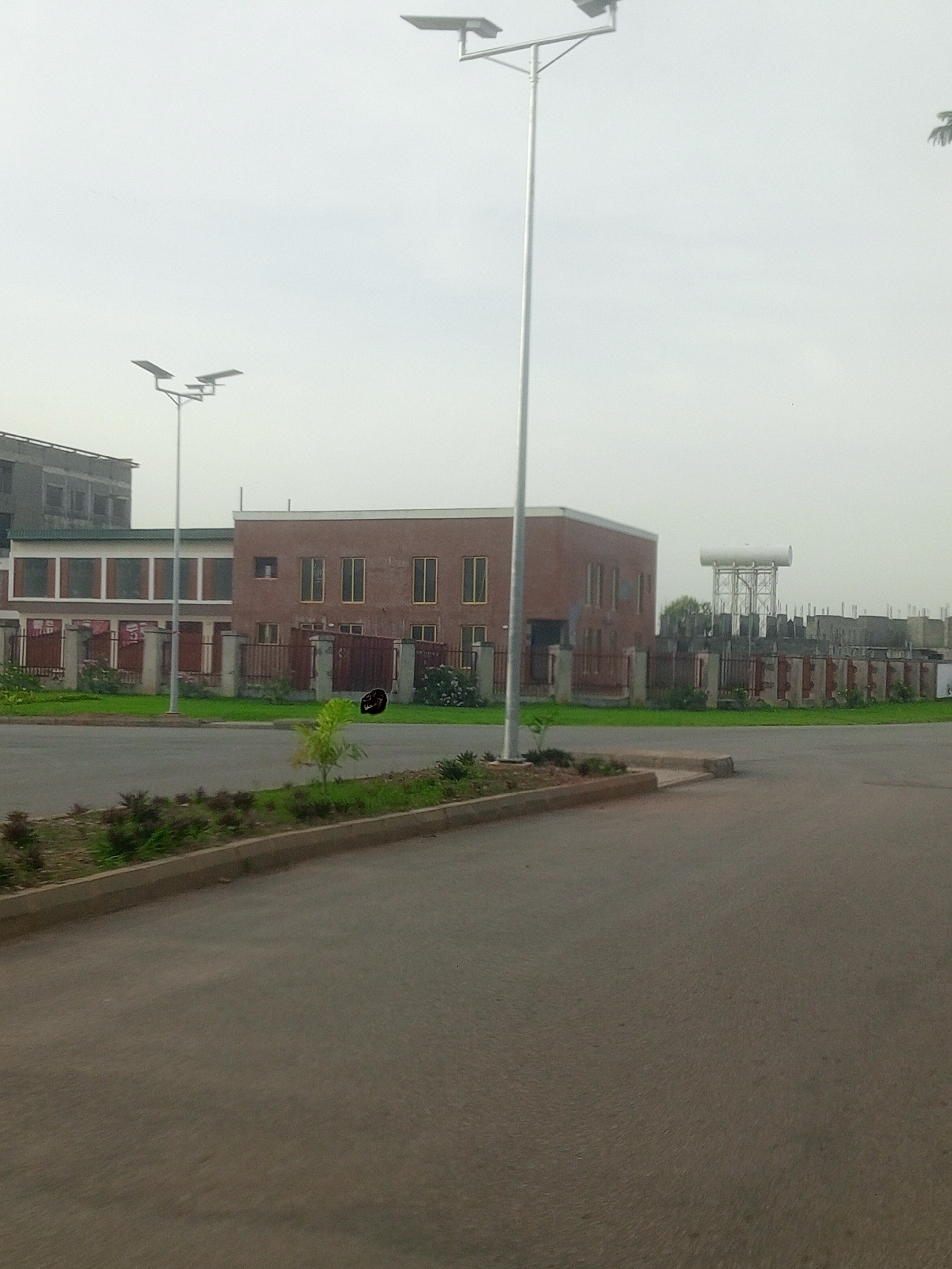 The image captures an overcast day in Gwarinpa, Nigeria, focusing on a street scene with modern architecture and infrastructure. In the foreground, a paved road occupies the lower half of the frame, bordered by a concrete curb and a strip of manicured greenery with small shrubs. A single modern street light with two lamps stands prominently in the mid-ground, to the left of center, its metal pole extending upwards against the pale sky.
Behind the street light and greenery, a cluster of buildings forms the backdrop. To the left, a multi-story building with dark facades and visible window frames suggests a commercial or residential complex. Adjacent to it, a smaller, reddish-brown brick building with a symmetrical facade and rows of evenly spaced windows is visible. This building is enclosed by a low wall with decorative concrete pillars and dark metal fencing, separating it from the street.
Further to the right, a construction site is evident. Partially built structures with exposed concrete pillars and beams are visible, topped by a large, white cylindrical water tank on a metal framework. This suggests ongoing development or expansion in the area. The overall impression is one of urban development with a blend of completed modern structures and ongoing construction, under a uniformly gray sky.