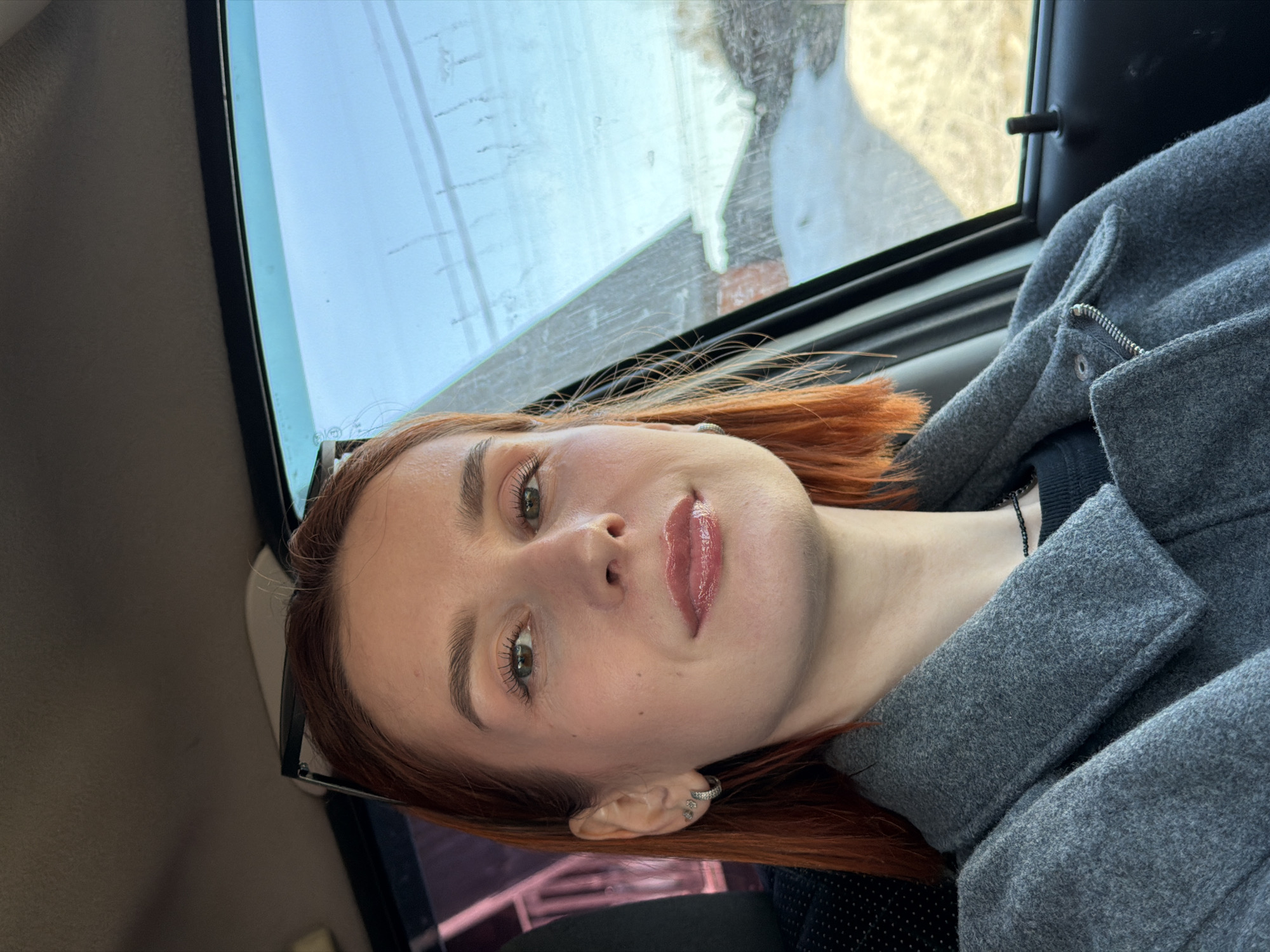 A red-haired woman is in the passenger seat of a car, facing the camera. She is wearing a gray wool coat and a black beaded necklace. The car window behind her reveals a blurry outdoor scene, possibly in Chaikovsky, Russia, due to the presence of houses and what appears to be snow or frost. The location cannot be positively identified as Chaikovsky, Russia, from the image alone.