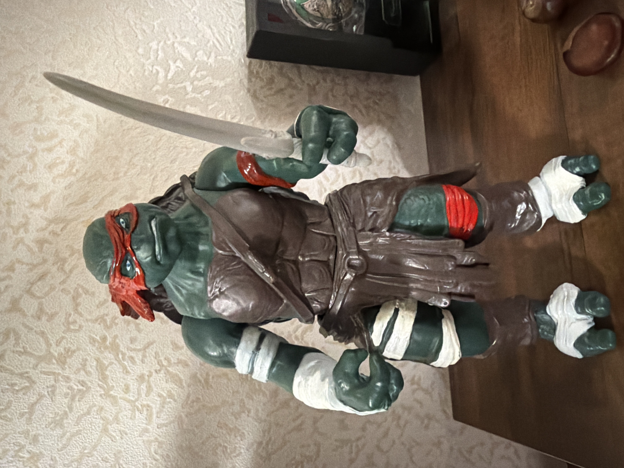 The image shows a detailed action figure of Raphael from the Teenage Mutant Ninja Turtles, standing on a wooden surface. The figure is green-skinned, with a red bandana over its eyes, and is wearing brown armor and a brown skirt-like garment. White bandages are wrapped around its arms and feet. Raphael is holding a silver sword in its right hand. In the background, there is textured wallpaper and a black rectangular object. The scene appears to be indoors, possibly on a shelf or table. The specific location of Yablonovskiy, Russia is not determinable from the visual information present in the image.