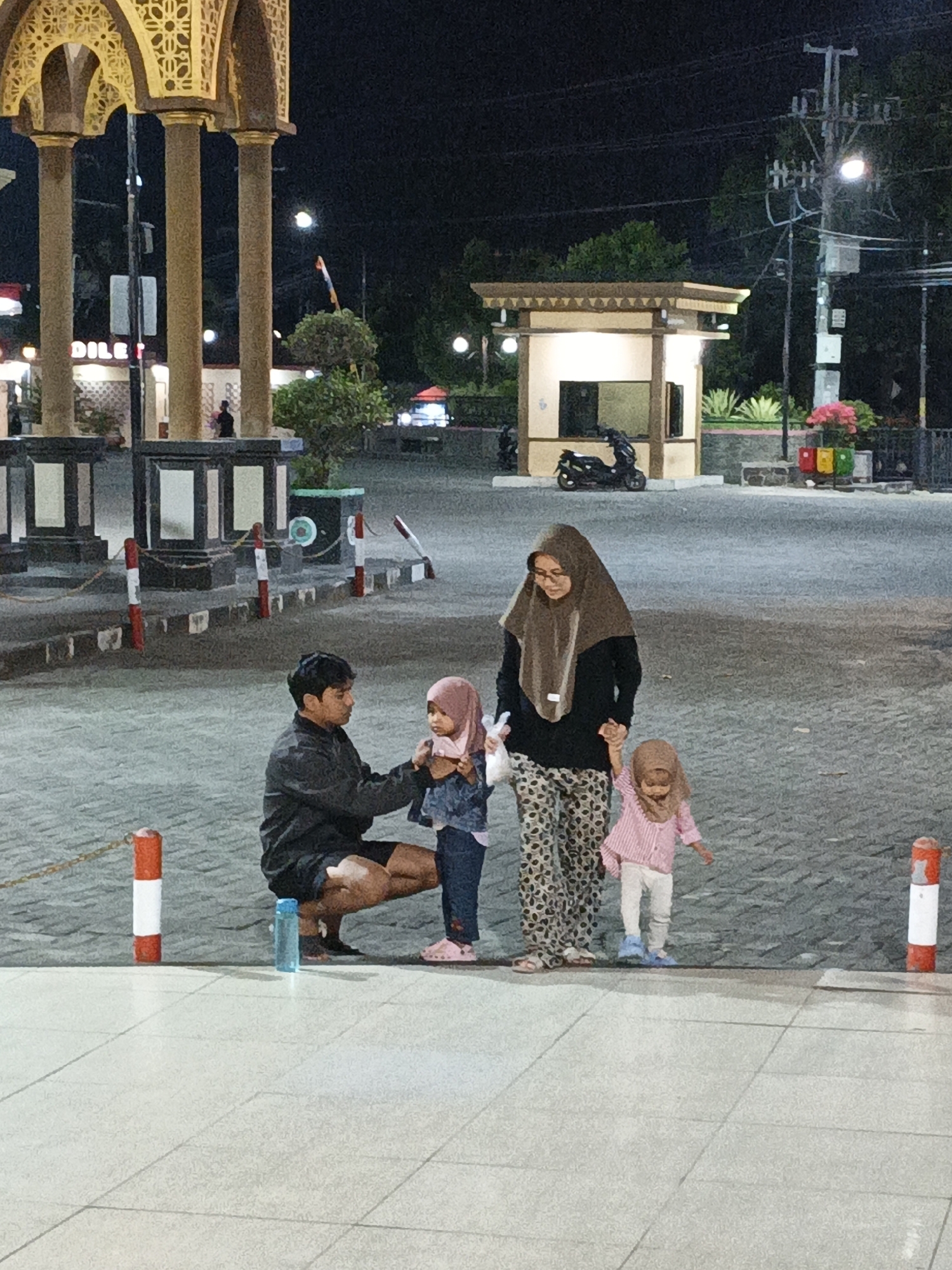 In Jonggrangan, Indonesia, at night, a family of four stands on a paved area in front of a building with a booth. A man is crouching down, interacting with a young girl wearing a pink hijab. A woman, also in a hijab, stands next to him, holding the hand of another young girl in a pink striped shirt. The background shows architectural elements, trees, and lighting.